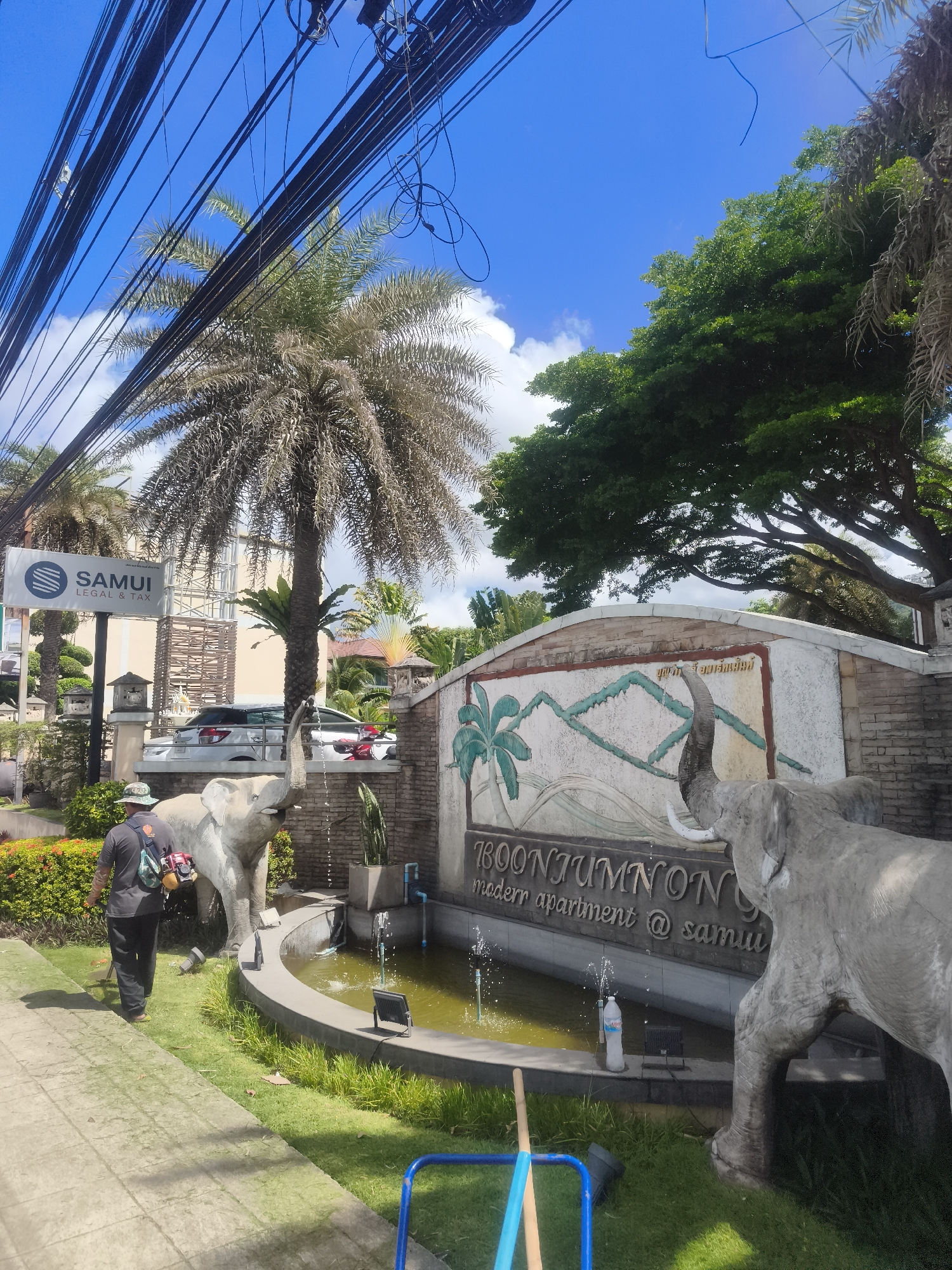In the city of Ko Samui District, Thailand, a man is using a string trimmer to cut grass near a decorative fountain. The fountain features two large elephant statues, one on each side, with water spraying from their trunks. The fountain is surrounded by a curved concrete wall with a decorative mural depicting mountains and palm trees. The mural also includes text that appears to read "BOONJUMNO" and "modern apartment @ samui". In the background, there is a building with a sign that says "SAMUI LEGAL & TAX" and lush green trees. The sky is blue with some clouds, and a tangle of electrical wires runs across the top of the image. The scene is illuminated by bright sunlight, indicating it is daytime.