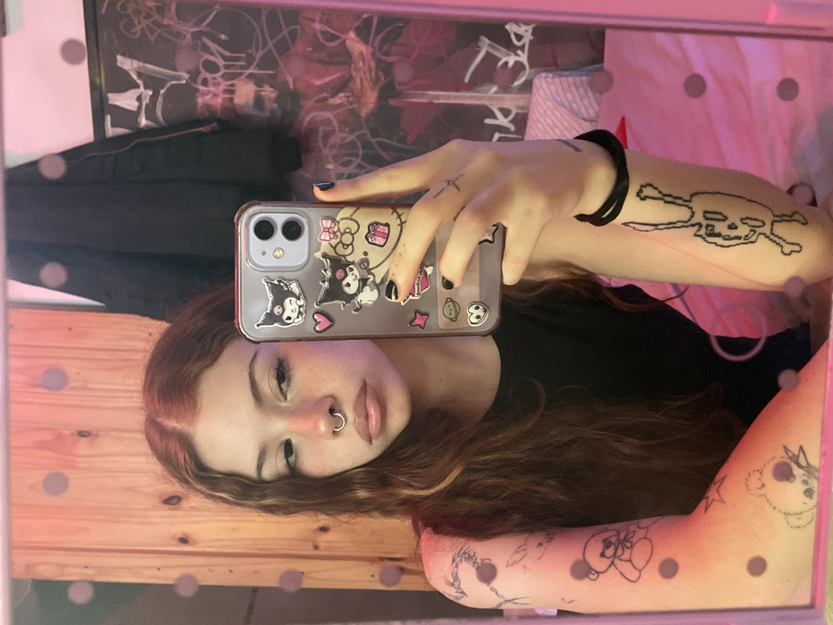 A young woman takes a selfie in a mirror. She has light skin, red hair, and a nose ring. Her phone case is decorated with Kuromi stickers. The woman's left arm displays several tattoos, including a skull and crossbones and a teddy bear. The background includes posters, clothes hanging on the wall, and what appears to be a wood-paneled wall at the bottom of the mirror. The mirror itself has a polka-dot pattern along the edge.