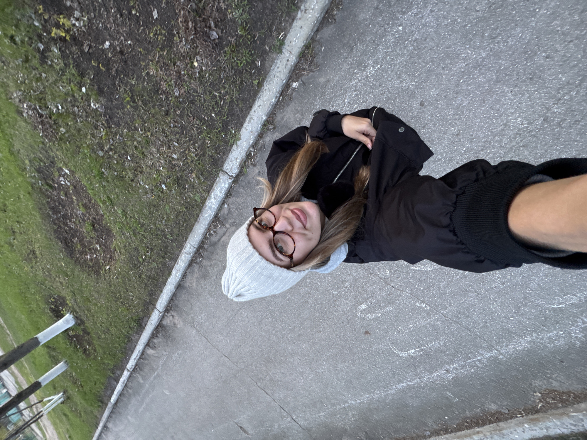 The image is a high-angle, tilted photograph, looking down at a young woman on a paved surface next to a grassy area. The woman, positioned in the center-left, is seen from approximately the waist up. She has shoulder-length, light brown hair and wears a light gray knitted beanie, brown-rimmed glasses, and a black jacket or coat. She is looking upwards towards the camera with a gentle, slight smile, her arms bent with hands held in front of her.
The ground she stands on is a gray, paved surface, likely concrete or asphalt, with faint white chalk markings visible on the right side. A concrete curb runs diagonally across the image, separating the paved area from an unkempt strip of grass and dirt. This grassy patch contains scattered dry leaves, small stones, and patches of bare earth, indicating a natural or less maintained outdoor environment. In the far background on the left, several metallic or gray posts are partially visible, suggesting a fence or other structure.
The activity appears to be the woman taking a high-angle selfie or having her photo taken from an elevated position. The overall lighting suggests it is daytime, possibly overcast or late afternoon, given the soft illumination and lack of harsh shadows. Her attire (beanie, jacket) suggests cool weather. The scene appears to be an outdoor urban or suburban setting, perhaps a park path or a sidewalk. Her expression conveys a sense of calm or pleasantness. No discernible text is visible in the image.
Location context: Unknown, Unknown