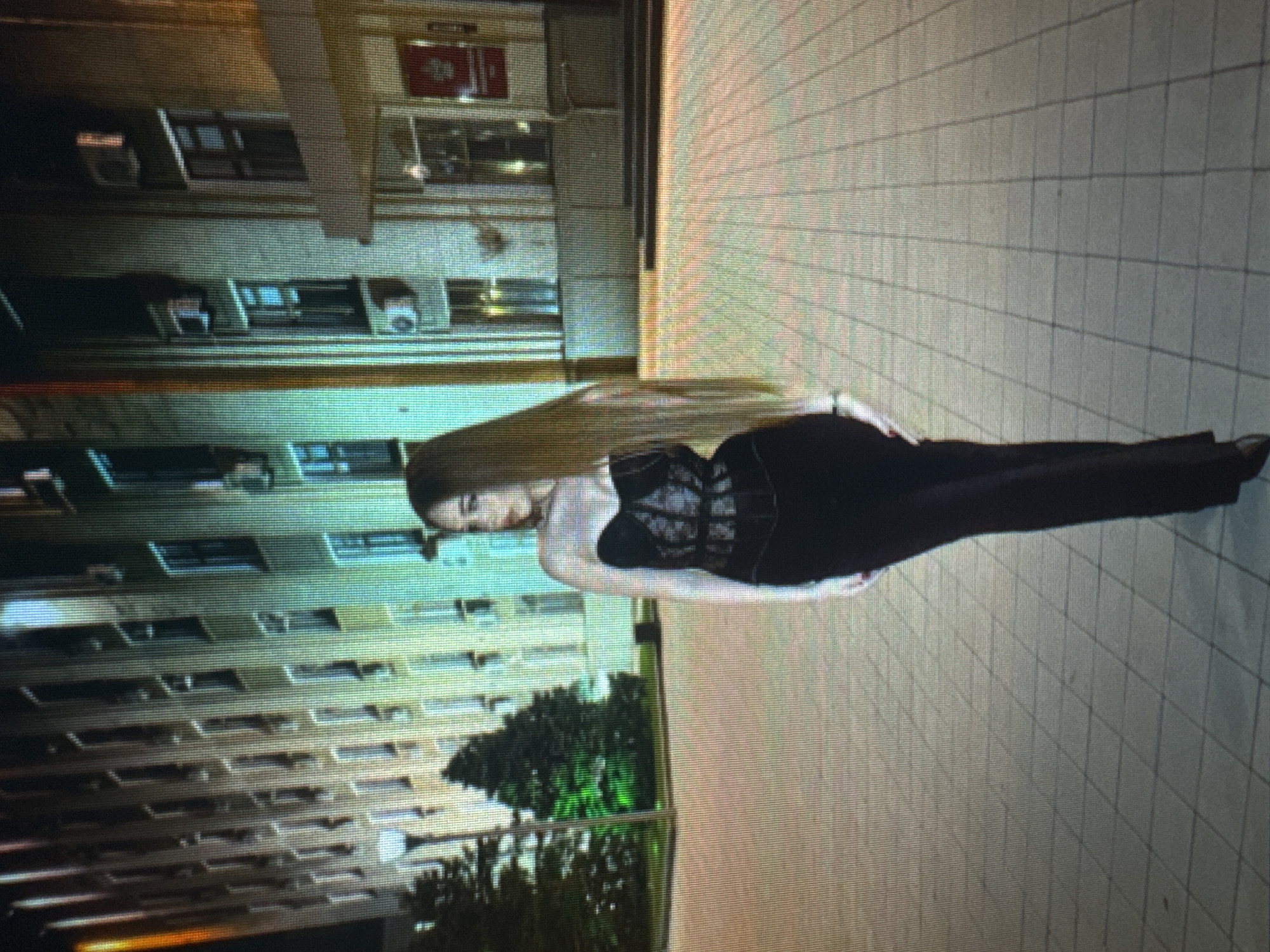 The media file depicts a woman standing on a tiled surface, possibly a sidewalk or plaza, in front of a building. The woman is wearing a black corset-like top and black pants, with her long blonde hair falling down her back. She is positioned in a way that suggests she is either posing for a photo or has just been photographed. The building behind her appears to be an apartment complex or office building with multiple windows. The time of day seems to be evening or night, as evidenced by the artificial lighting present in the scene. There are no clear indications of weather conditions. The setting could be urban, with the tiled ground and multi-story building. The image is slightly pixelated, making it difficult to discern finer details. No visible text is present in the image.