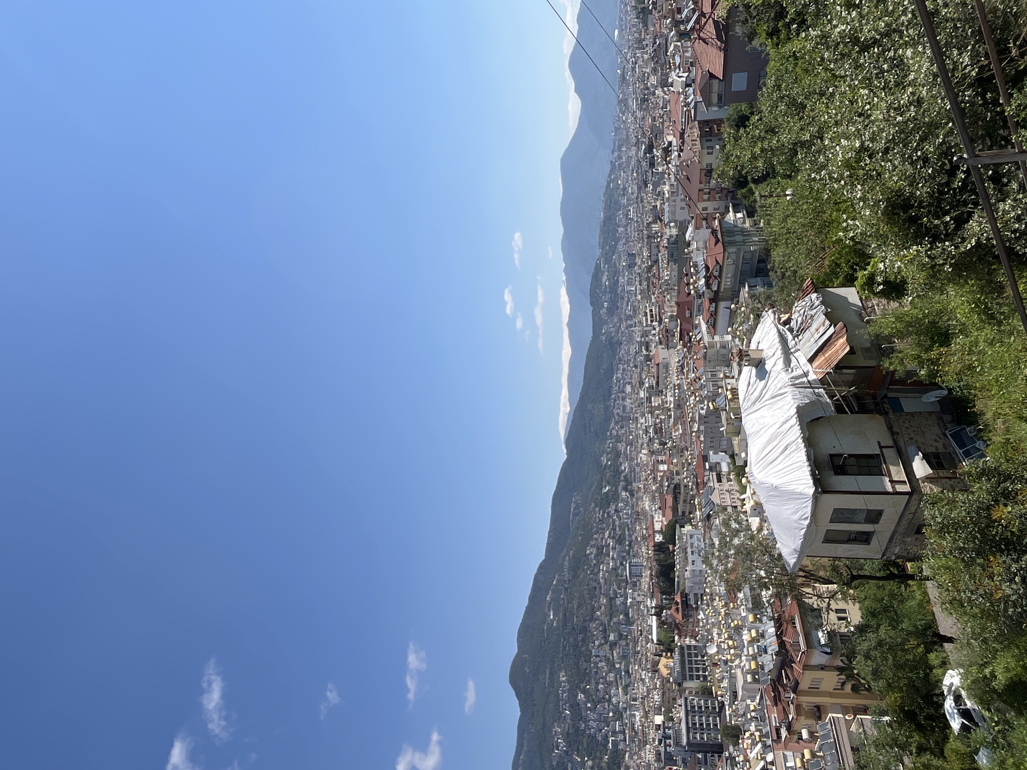 The image shows an elevated view of a densely populated area, likely the city of Alanya, Turkey, given the architectural style and geography. The scene is dominated by closely packed buildings, ranging in height from low-rise to mid-rise structures, intermingled with trees and patches of greenery. Mountains are visible in the background under a clear, blue sky with a few scattered clouds. The image appears to be taken from a high vantage point, possibly from a cable car or similar elevated structure, as evidenced by the presence of cables in the upper right corner. The focus is on the built environment and the natural landscape that surrounds it.
