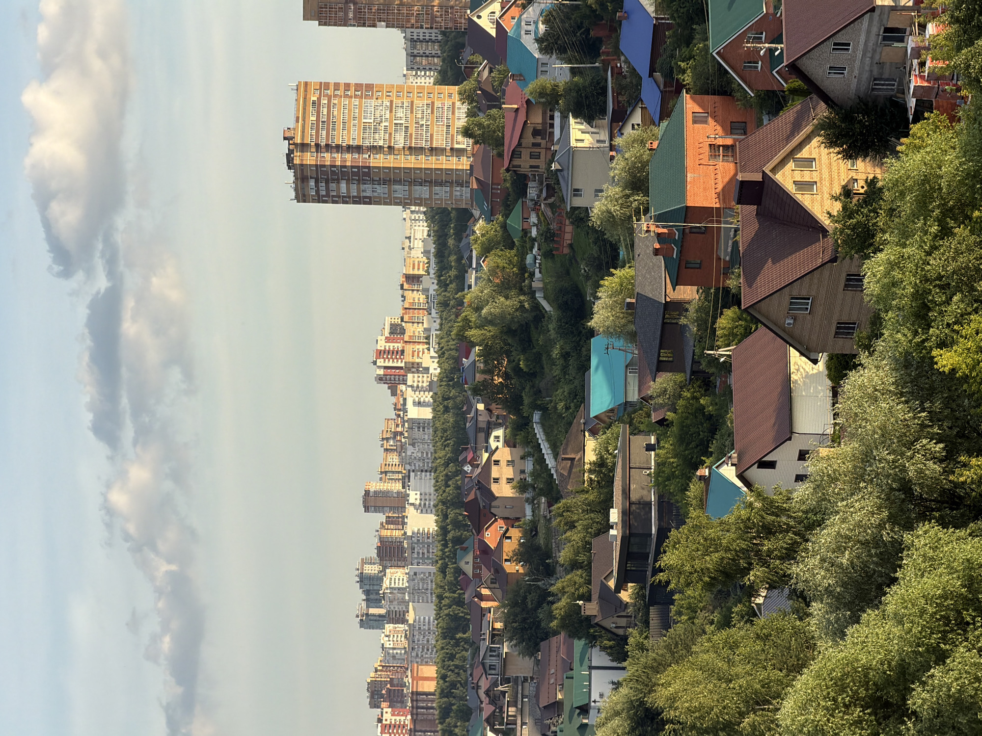 This is a photograph of a cityscape in Kazan, Russia, taken on a clear day. The image shows a mix of residential and high-rise buildings, with dense green trees interspersed throughout the scene. The buildings vary in architectural style and color, with some featuring bright, colorful roofs. The time of day appears to be late afternoon, as indicated by the lighting. There is no visible text in the image. The overall impression is one of a well-developed urban area surrounded by abundant greenery, suggesting a pleasant and possibly residential district.