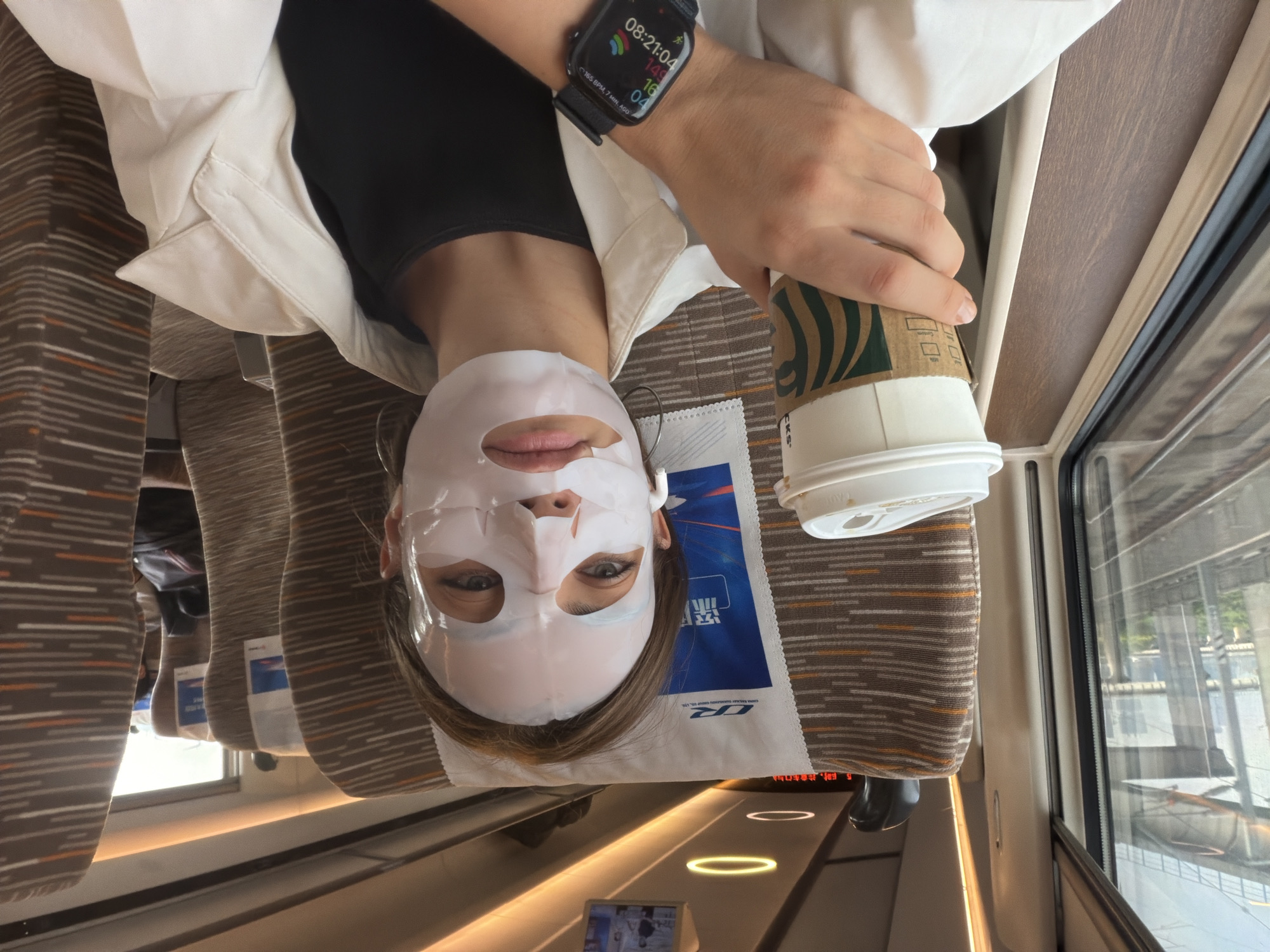 A young woman with a white sheet mask covering her face sits on a train, holding a Starbucks cup in her right hand. She is wearing a white open jacket over a black top and a black smartwatch on her left wrist. She has on small hoop earrings. The train seats are upholstered in a brown and white striped fabric. Through the window, train tracks and a bridge are visible, suggesting the train is moving through an urban or suburban area. The lighting on the train appears to be artificial, with overhead lights. The location is identified as Panyu District, China.