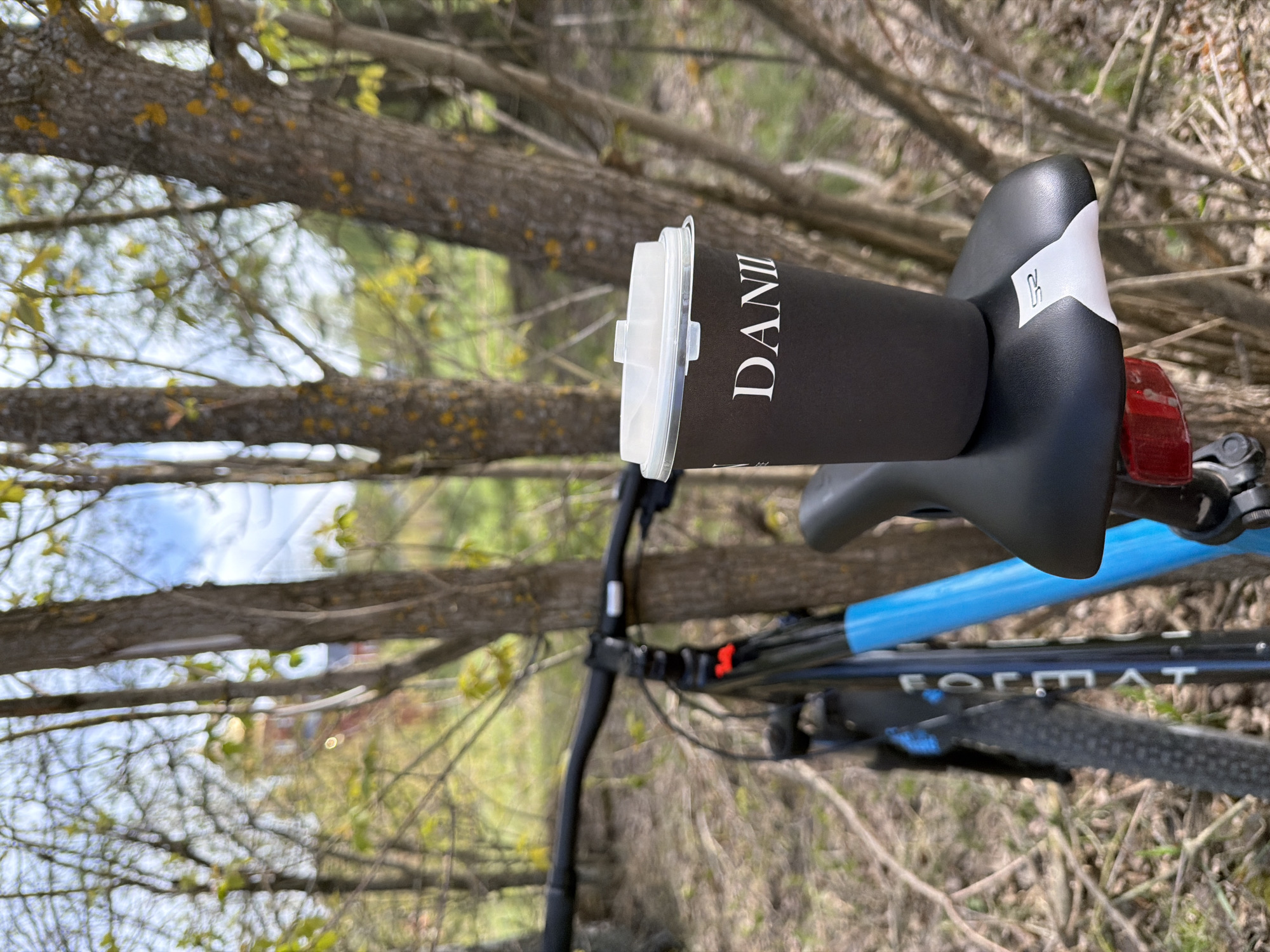 A black coffee cup with a white lid, labeled "DANI," sits precariously on the black and white bicycle seat. The bicycle, partially visible, is blue and black with the word "FORMAT" printed on the frame. The bicycle is parked outdoors amidst trees and grass, possibly in a park or wooded area. The background is blurred, but glimpses of the sky and some buildings are visible through the trees. The image is taken in daylight. It's difficult to pinpoint the precise location within Chaikovsky, Russia, based solely on the visual details.
