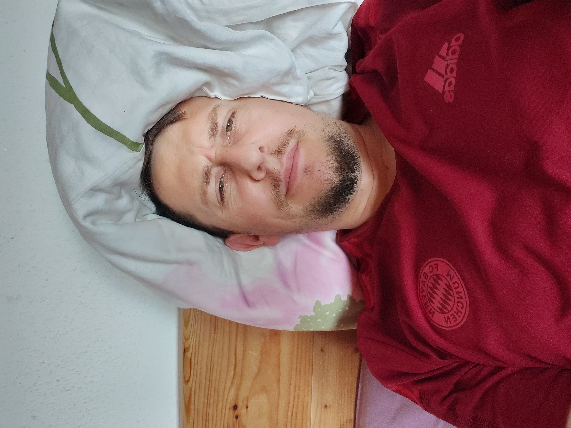 An adult male is pictured lying in a bed, looking directly at the camera. He has dark hair, a beard, and a neutral expression with a slight furrow in his brow. He is wearing a dark red short-sleeved shirt featuring the mirrored FC Bayern München crest on the left chest and a mirrored Adidas logo on the right chest, indicative of a front-facing camera shot. His head rests on a pillow with faint pink and green patterns, partially covered by a white duvet displaying a prominent green linear design. Behind him is a light wooden headboard and an off-white, textured wall. The setting is an indoor bedroom. No visual information within the image provides specific cues to confirm a location in the city of Straubing, Germany.