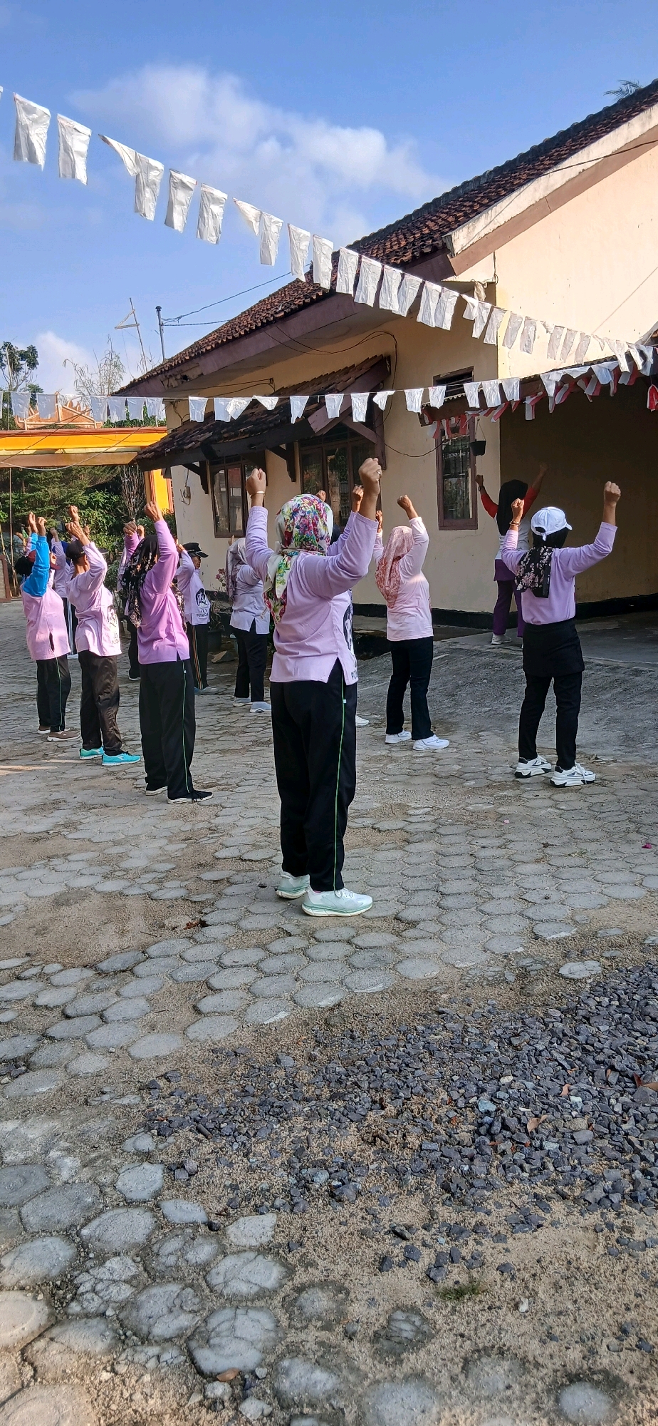 The image captures a group of women engaged in an outdoor exercise activity, likely a form of aerobics or dance, on a paved courtyard in front of a building. The women are dressed in coordinated pastel-colored shirts and dark pants, with some wearing headscarves. Their arms are raised in unison, suggesting they are following an instructor or a routine. The building in the background appears to be a single-story structure with a tiled roof and several windows. White triangular flags are strung across the scene, possibly for a celebration or event. The ground is a mixture of interlocking paving stones and some gravelly, sandy areas, indicating it could be a public space, schoolyard, or community gathering area. The lighting suggests it is daytime, with a clear blue sky overhead. The scene is likely set in the city of Metro, Indonesia, as the flags might be red and white, signifying the national colors of Indonesia, often displayed during national holidays or celebrations.
