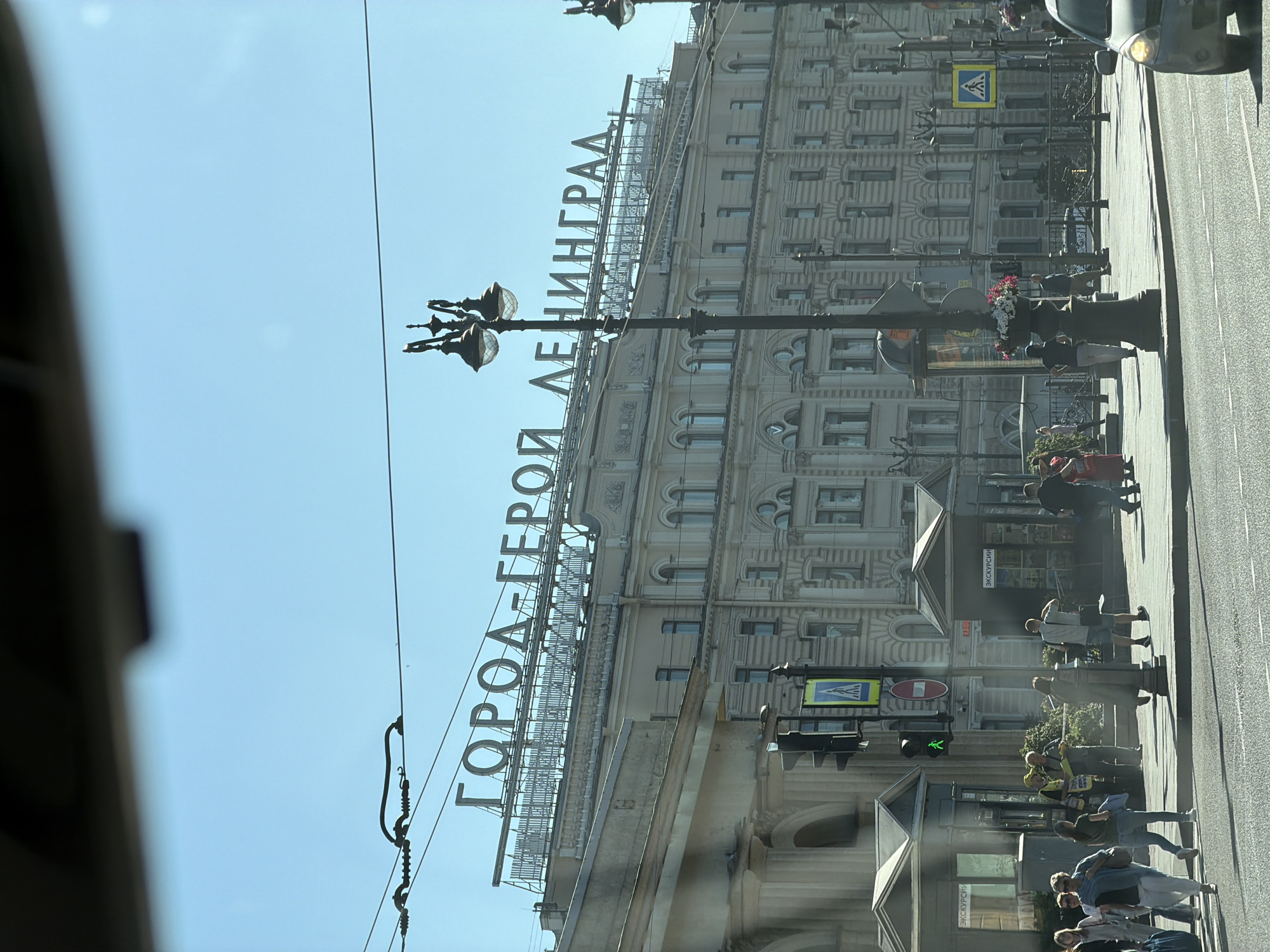 The image shows a street scene in Saint Petersburg, Russia, with a large building displaying the Cyrillic text "ГОРОД-ГЕРОЙ ЛЕНИНГРАД" (Hero City Leningrad) prominently on its facade. The building appears to be an architectural landmark. In the foreground, people are walking on the sidewalk and crossing the street. Traffic lights and street lamps are visible, indicating an urban environment. A car is also present in the right side of the frame. The sky is clear and blue, suggesting a sunny day. The overall impression is that of a bustling city street on a bright day.