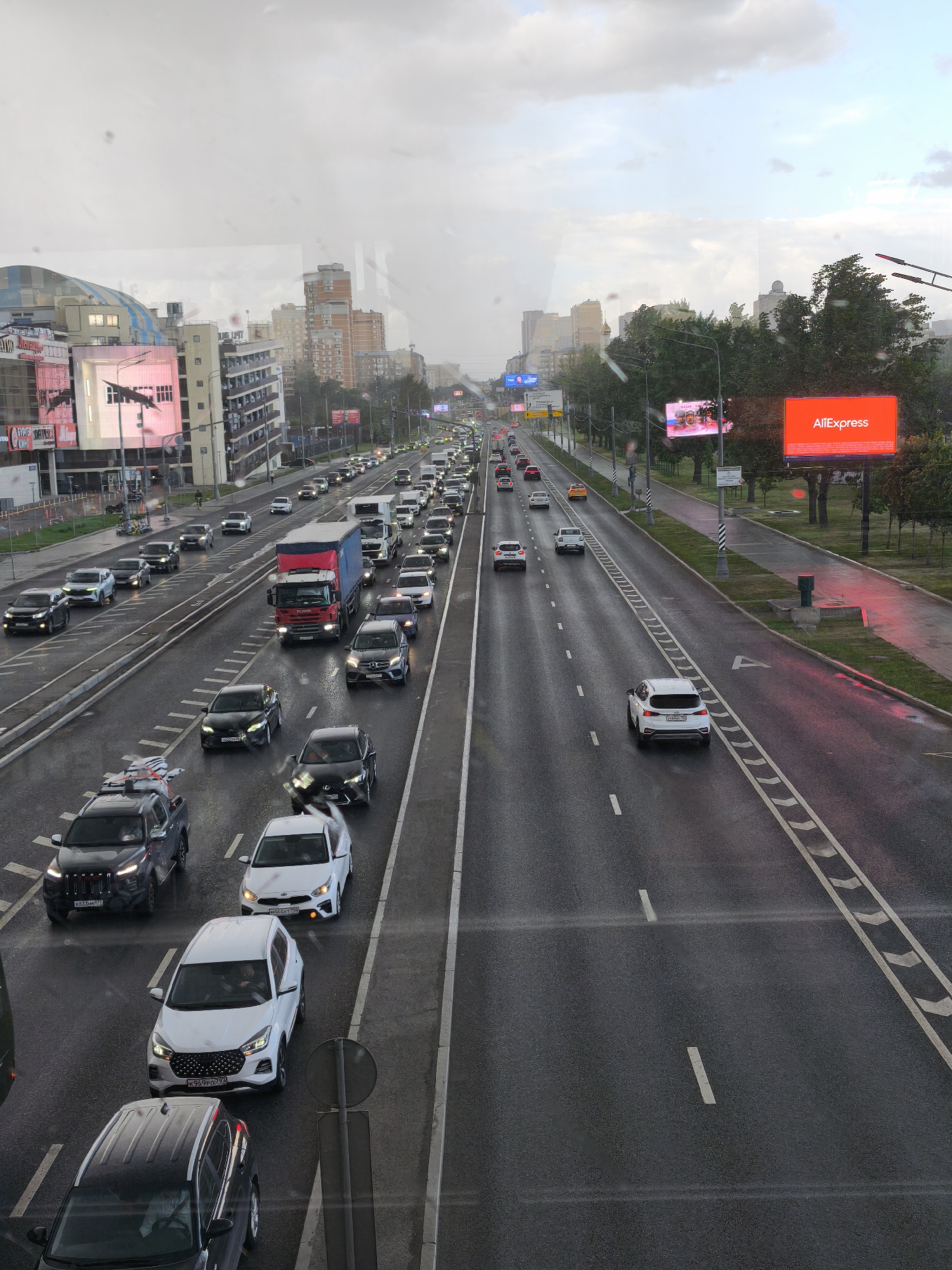 The image shows a busy multi-lane road in Moscow, Russia. It appears to be taken from an elevated position, possibly a bridge or building. The road is wet, suggesting recent rain. There is heavy traffic in both directions, consisting of cars, trucks, and other vehicles. Buildings and billboards line the road, and there are green spaces on the side with trees and grass. An "AliExpress" billboard is prominently visible. The sky is overcast, contributing to the grey and rainy ambiance of the scene.