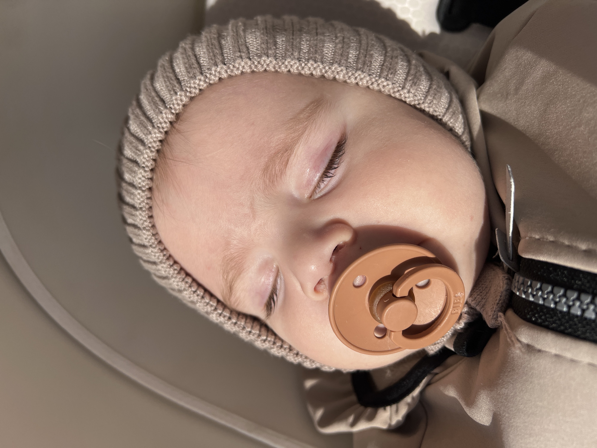 A close-up, slightly angled overhead shot shows a sleeping baby with a pacifier in its mouth. The baby is wearing a knitted beige cap. The baby is wearing a light beige jacket with a black zipper. The background is a neutral, light color, suggesting the baby is in a car seat or similar carrier. The image does not provide any information regarding a city.