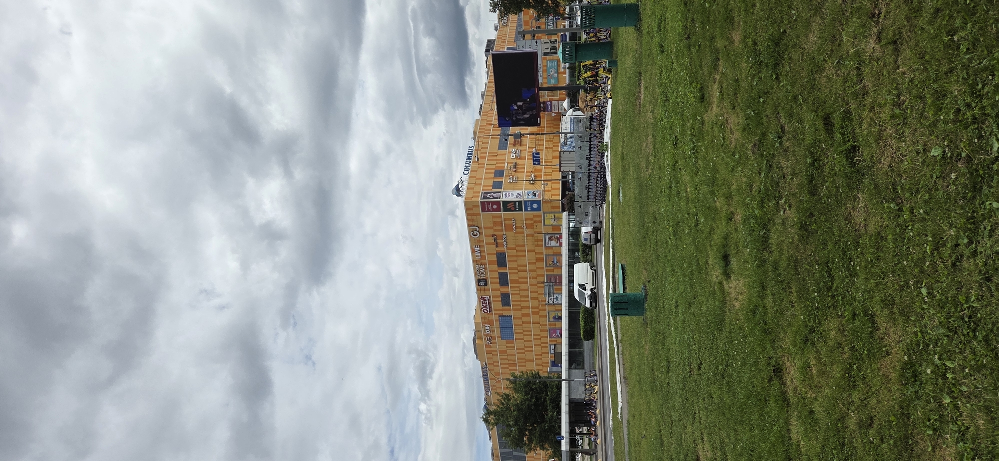 The image captures a wide shot of the Columbus shopping center in Moscow, Russia, on a cloudy day. The large, orange building is the dominant feature, with various signs for stores like "COLUMBUS," "RE CLH," "OKEY," "askona," "HOME," "LIME," and "GJ" visible on its facade. A large digital billboard is also mounted on the building's exterior.
In front of the shopping center, a row of parked electric scooters is visible, and further to the left, a bike rack with several bicycles. Two white vans are parked on the road in front of the building. A patch of green grass is in the foreground, with two green trash cans. The sky is overcast with thick, grey clouds. A tree is located to the left of the shopping center. The overall scene depicts an urban environment with commercial activity.