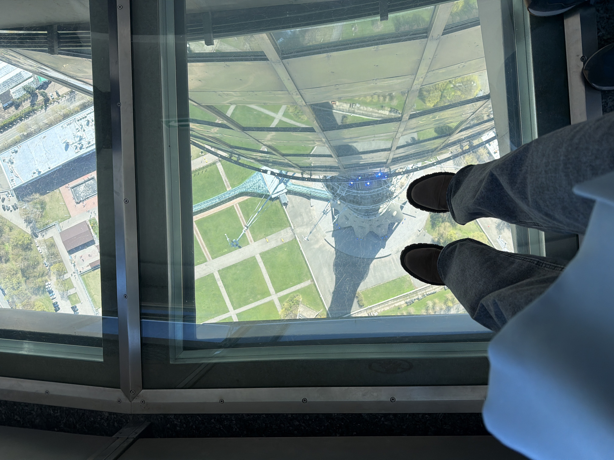 The image, taken from a high vantage point, likely a tower or tall building, features a person standing on a glass floor. The viewer's perspective is directly down, looking through the glass to the ground below. The person is wearing grey jeans and brown shoes. Their shadow is also cast on the area below. The area below contains the base of the tower, a lawn, and some buildings. Although a location in Khimki, Russia was requested, the image doesn't give enough details to provide that information.