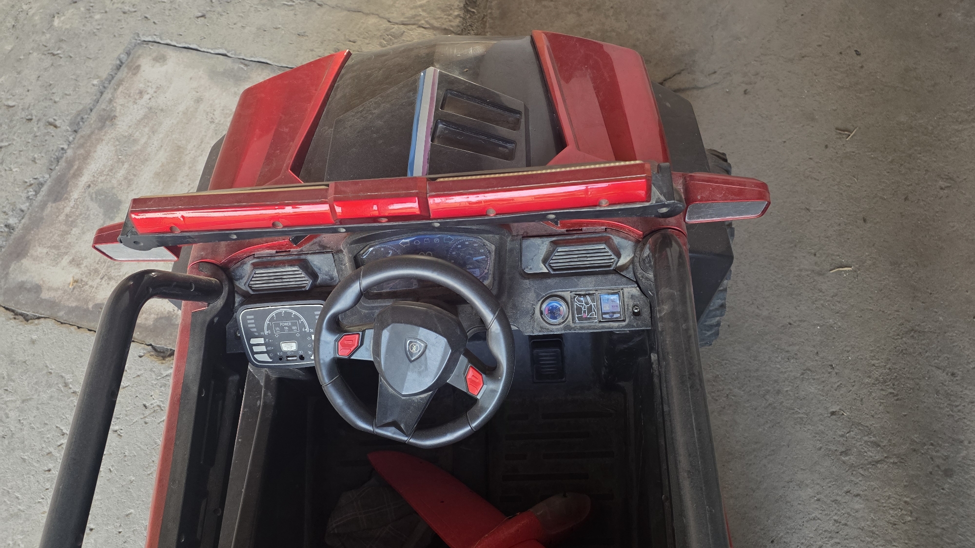 An overhead shot shows the interior of a red toy electric car. The car is parked on a concrete surface, which appears to be a sidewalk or driveway. The car's dashboard features a steering wheel, a speedometer, and various buttons and controls. A red toy airplane lies on the passenger side floor. The environment appears to be outdoors, possibly in a residential area. The location is identified as Chuy, Kyrgyzstan.