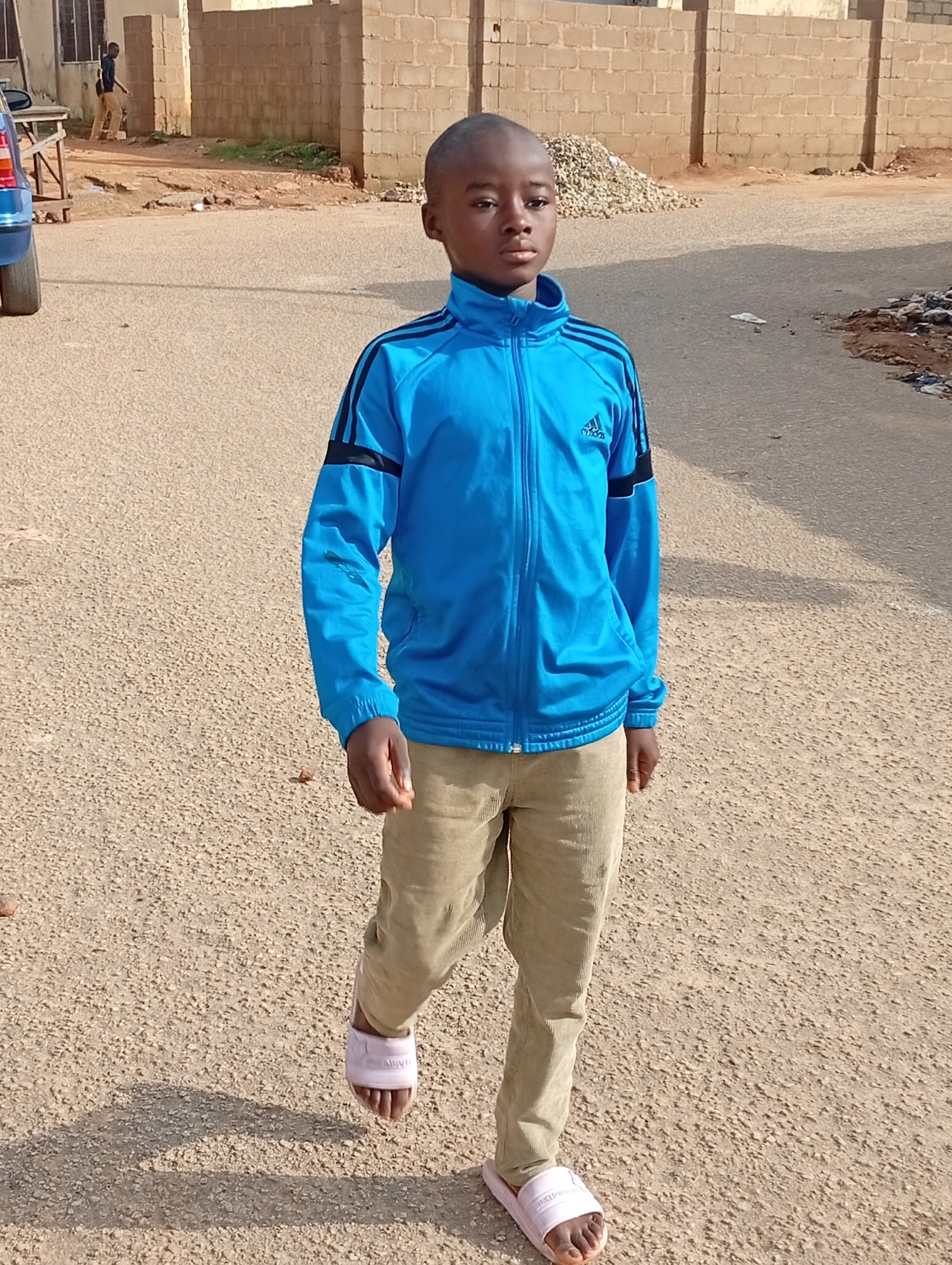 A young boy stands in the middle of a paved road, looking towards the camera. He is wearing a bright blue track jacket with black stripes on the sleeves, light brown corduroy pants, and pink slides. The boy has short, dark hair and a serious expression. The background shows a brick wall and some construction materials, suggesting an urban or suburban environment. The road appears to be a typical street with a rough, gravelly surface. The lighting indicates it is daytime, with shadows cast by the boy and objects in the background.