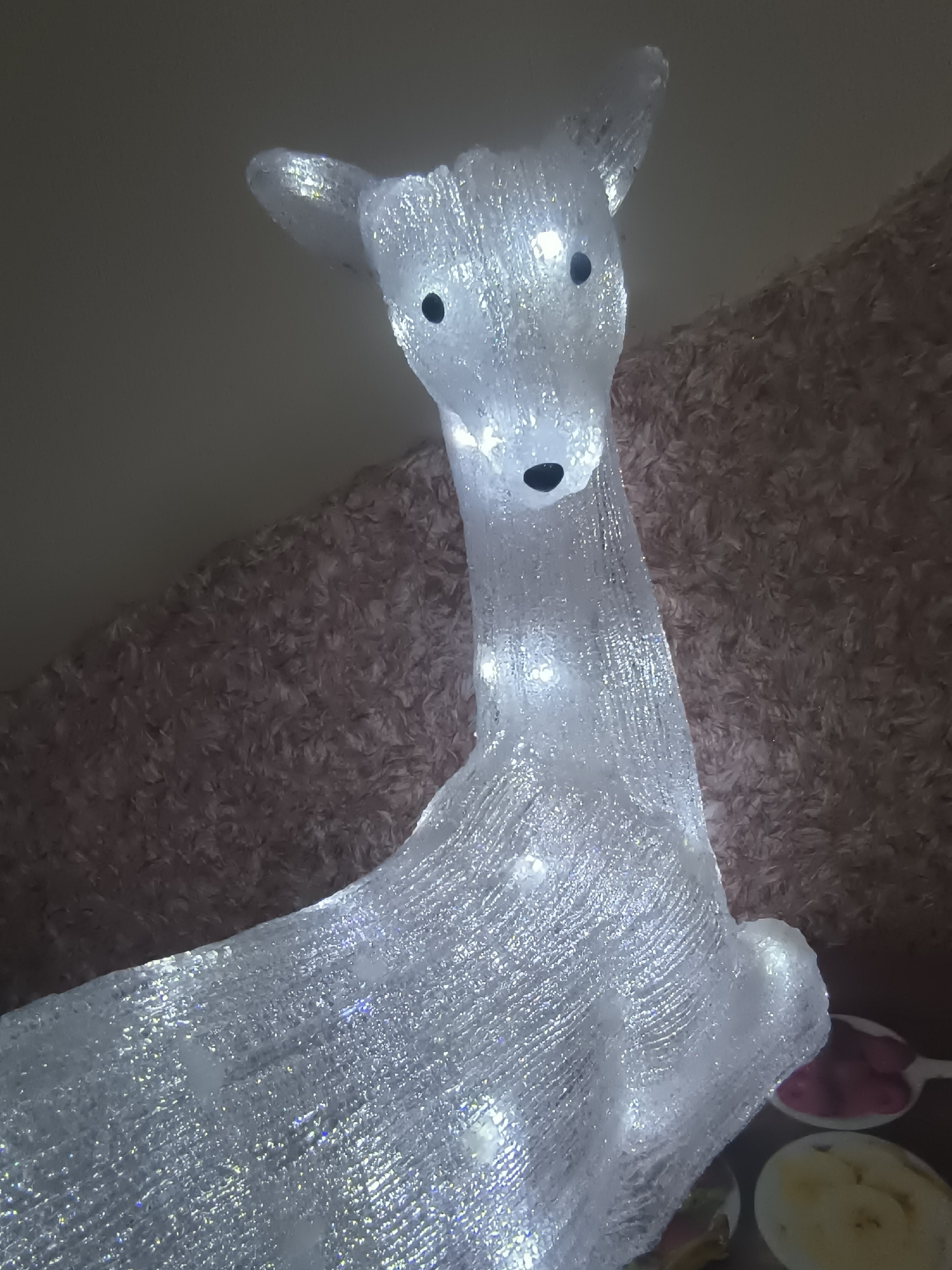 A close-up, low-angle indoor shot shows a lit-up reindeer Christmas decoration. The decoration is made of clear, sparkly material and illuminated by white lights, giving it a translucent appearance. It is positioned in front of a textured, furry, pale pink background. To the right and slightly out of focus, there are several small dishes containing food items, including what appear to be berries and sliced fruit. The scene is likely a domestic setting, possibly decorated for the holiday season. The location in Klaipėda, Lithuania, is not discernible from the visual information provided.