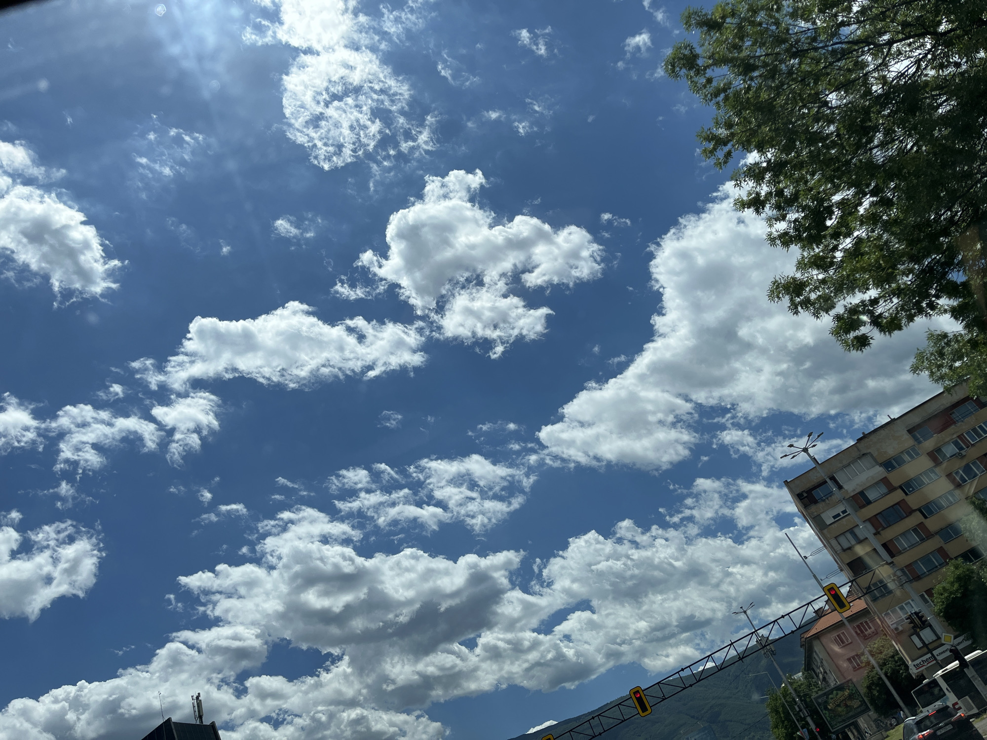 This is a vertical, medium shot of a blue sky filled with scattered white cumulus clouds. The sun is visible in the upper right quadrant, creating lens flare. In the foreground, the tops of leafy green trees are visible on the right. In the bottom left quadrant, a section of a city appears, with buildings and streetlights. One building is a light brown, multi-story structure with many windows. Another building behind it is darker and more recessed. A black metal pole with yellow traffic signals is also visible on the left. A dark-colored vehicle is partially visible at the bottom left. The scene appears to be outdoors in an urban environment, possibly during the daytime.