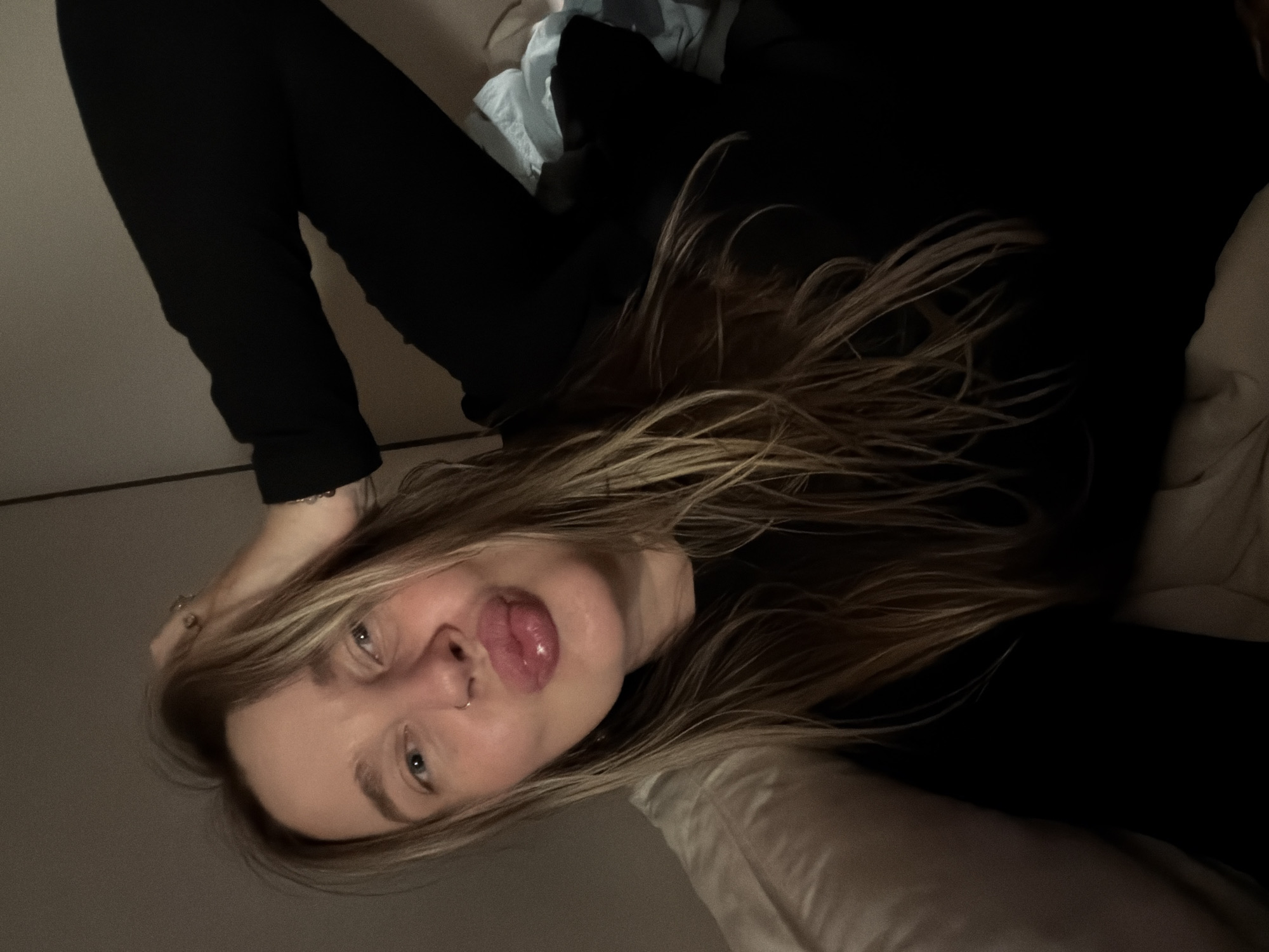 A woman with long blonde and brown hair is lying down. She is wearing a black long-sleeved shirt and has a nose ring. Her head rests on a white pillow, and her hair is spread around her. The background includes a white wall and some light-colored fabric. The image provides no information to indicate a location in Moscow, Russia.
