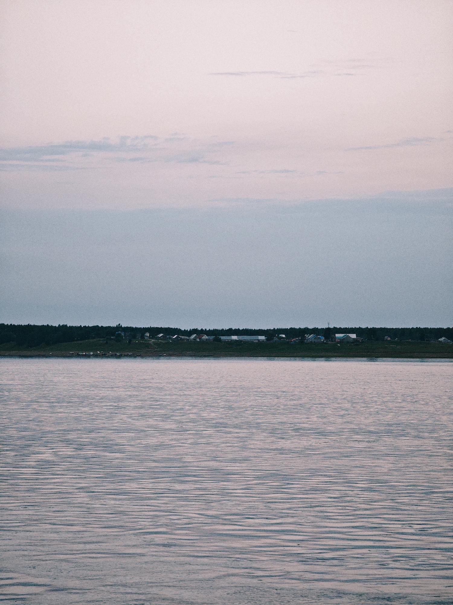 The image depicts a serene landscape in Nikulino, Russia, characterized by a vast expanse of calm water in the foreground and a tree-lined shore with a settlement in the distance. The water exhibits gentle ripples, reflecting the soft, diffused light of the sky. A dark, dense forest forms a backdrop to a small village, with houses and their roofs clearly visible, suggesting a remote, rural setting. The sky is a pale, hazy hue, with subtle cloud formations adding texture. The overall impression is one of tranquility and stillness, indicative of a quiet evening or early morning.