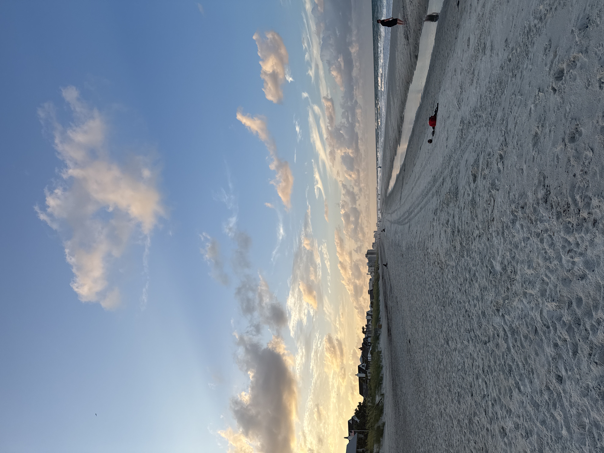 This is a photograph of a beach in Jacksonville Beach, Florida, United States of America, at dusk. The sand is light gray and appears somewhat wet near the water's edge. There are tire tracks on the sand, suggesting vehicle use. A small stream of water runs along the beach. In the distance, there are several houses and taller buildings visible along the coastline.
Several people are present on the beach. On the right side of the image, a person wearing dark shorts and a dark top stands facing the ocean. Further down the beach, other individuals are walking or sitting. In the middle ground, two red objects, possibly bags or towels, lie on the sand near the stream.
The sky is a gradient of blue and orange, with scattered clouds tinged with yellow and pink from the setting sun. A bird is visible in the upper left corner of the image. The ocean waves are breaking on the shore, creating a white foam line. The overall scene suggests a peaceful, early evening atmosphere at the beach.