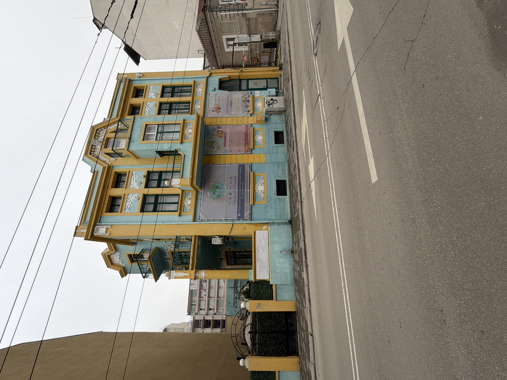 The image captures a colorful building in Varna, Bulgaria, identified as the "Happy Planet" private kindergarten, located next to a road. The building is painted in blue and yellow with decorative elements and multiple posters displayed on its facade. The road has lane markings and an arrow indicating traffic direction. Overhead power lines are visible against a gray sky. Neighboring buildings of varying heights and architectural styles flank the kindergarten, adding to the urban environment.