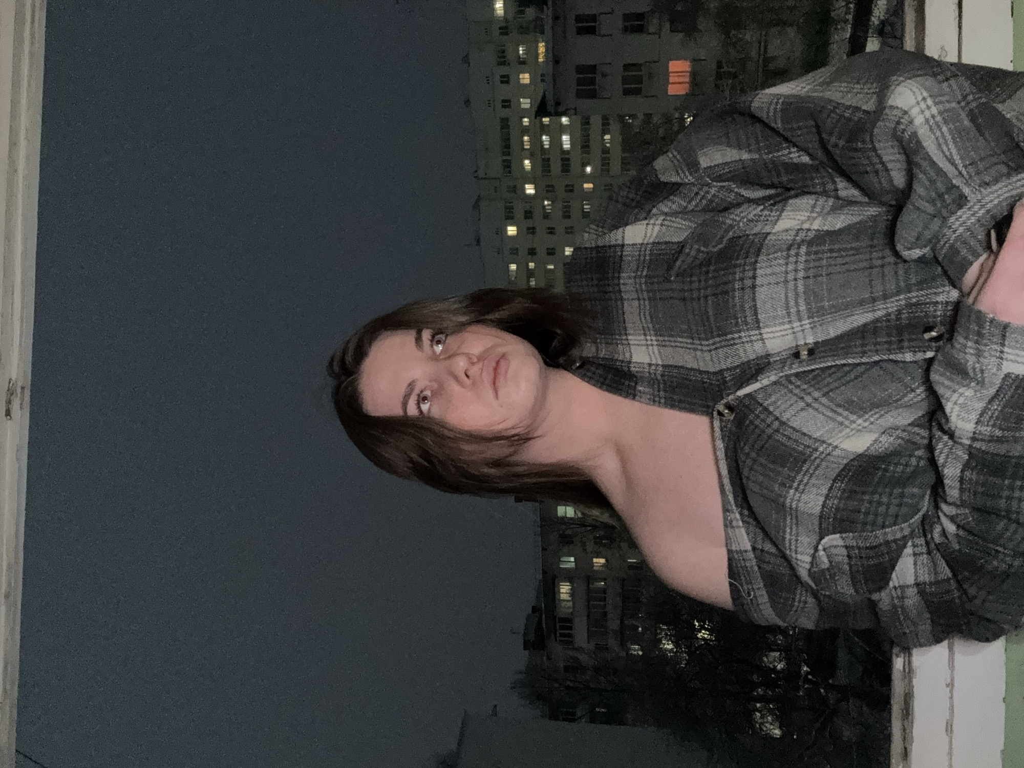 A young woman with short brown hair stands by a window in what appears to be an apartment building. She is wearing a gray and black plaid flannel shirt draped loosely over her shoulders. The lighting suggests it is either night or dusk.
Outside the window, the view encompasses other high-rise apartment buildings, many with lit windows. The overall atmosphere is urban.
While it is not possible to independently confirm if this photograph was taken in the city of Chaikovsky, Russia, the setting is consistent with the appearance of urban residential areas in the Russian Federation.