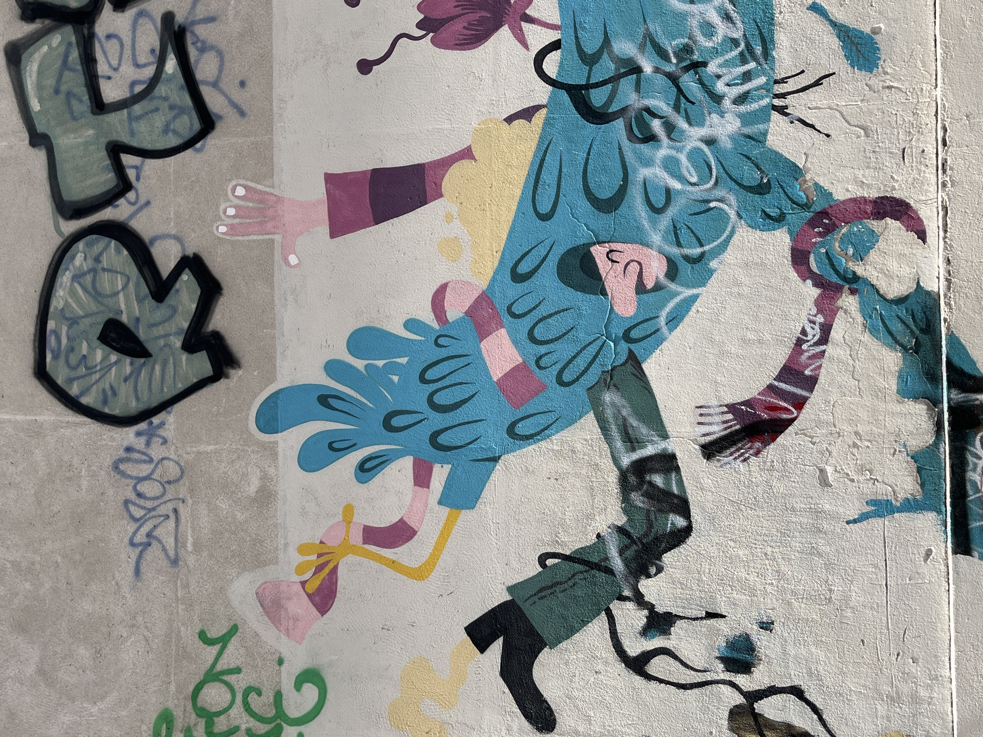 The image depicts a section of a wall featuring graffiti art, likely located in Paris, France, as suggested by the presence of various tags and urban art styles common in the city. The focal point of the artwork is a cartoonish blue creature with large, expressive eyes and an open mouth, rendered in a dynamic, almost falling pose. The creature has a pink face and is depicted with yellow limbs and a pink and purple striped torso.
Other elements of the graffiti include abstract shapes and lettering in various colors, including green, black, and a muted teal. There's a pink object held by one of the creature's yellow hands, appearing to be a cup or a similar vessel. Yellow wisps of smoke or liquid emanate from the creature's body and arm. A purple, flower-like motif is visible to the right of the main figure, with thin black stems. The background is a textured, off-white concrete wall, showing signs of wear and tear. Visible lettering, potentially tags, appears in blue and black over the concrete surface. The overall impression is that of a vibrant, albeit somewhat chaotic, urban art installation.