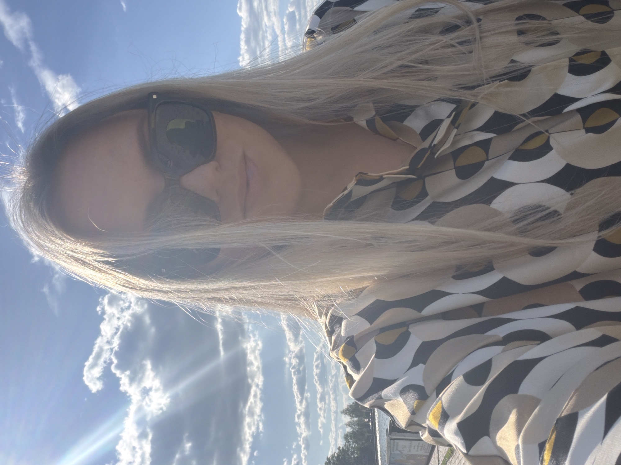 A blonde-haired woman wearing dark sunglasses and a patterned shirt is depicted in a selfie taken outdoors on a sunny day in the city of Votkinsk, Russia. Her long, straight hair falls over her shoulders. The shirt features a geometric pattern in cream, beige, dark gray, and yellow-gold hues. The background displays a bright blue sky with scattered cumulus clouds and visible sun rays emanating from the upper left. A body of water, likely a lake or river, with a tree-lined shore, is visible in the distance. A concrete or stone structure is partially observable on the left side of the frame.