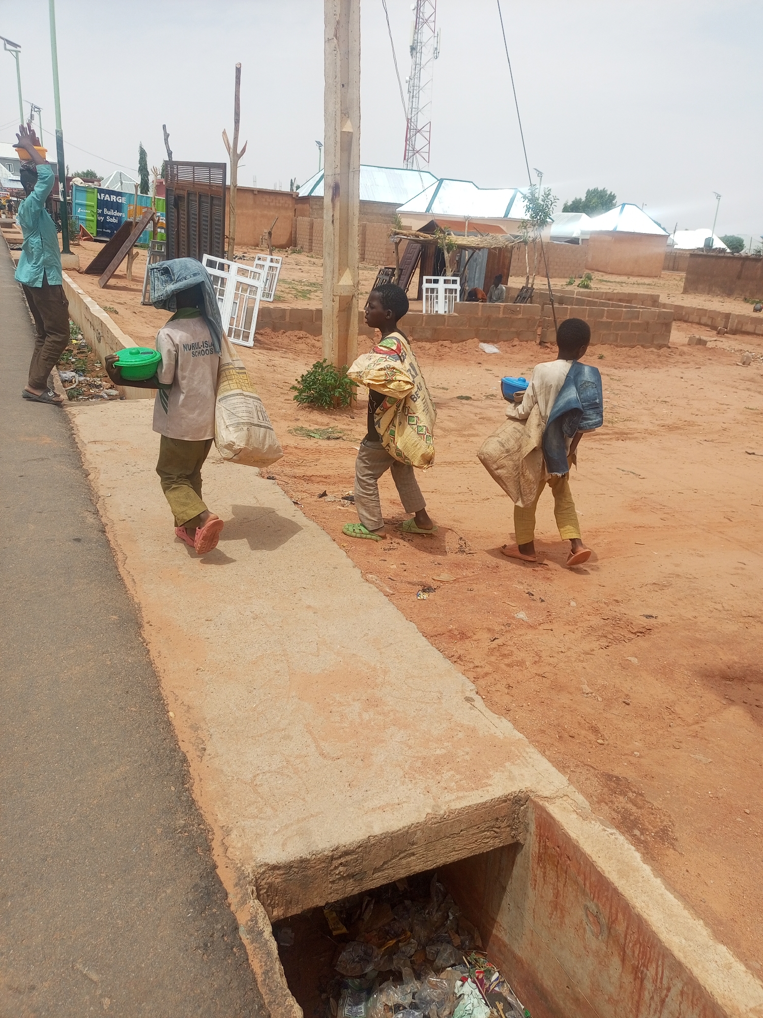Three children walk on a concrete sidewalk adjacent to a street. Each child carries a bag, and at least two carry additional items. An adult male stands at the edge of the road with one arm raised. The background shows a red, sandy terrain with low-rise buildings, some with corrugated metal roofs, and a cell tower. A concrete drainage ditch filled with trash runs alongside the sidewalk. The child nearest the camera wears a shirt with "NURUL-ISLA SCHOOLS" printed on the back. A sign with "LAFARGE" is visible in the background. The location is in Tempure, Nigeria.
