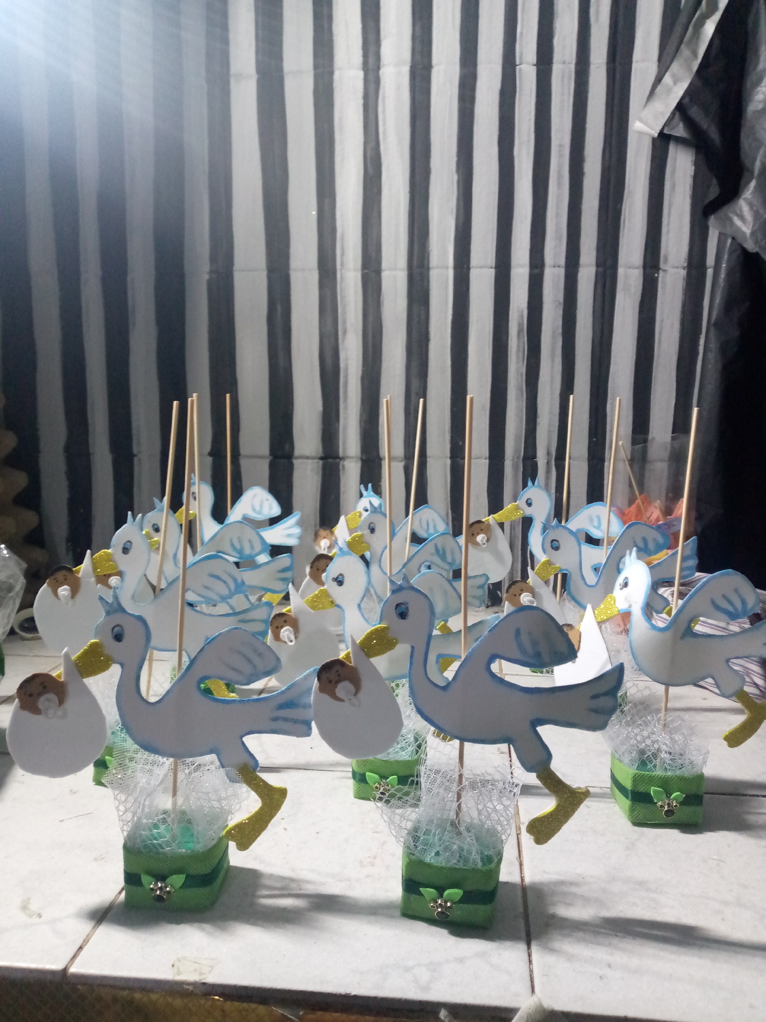 A collection of decorative stork centerpieces are arranged on a tiled surface. The storks are predominantly white and blue, with yellow beaks and legs, each carrying a baby figure in a white sack. They stand atop green, cube-shaped bases decorated with small leaves. A backdrop of black and white vertical stripes is visible, and there are wooden sticks emerging from some of the storks, likely for stabilization. The setting is indoors, potentially related to a baby shower or celebration. It is not possible to determine the location of Juan Griego, Venezuela.