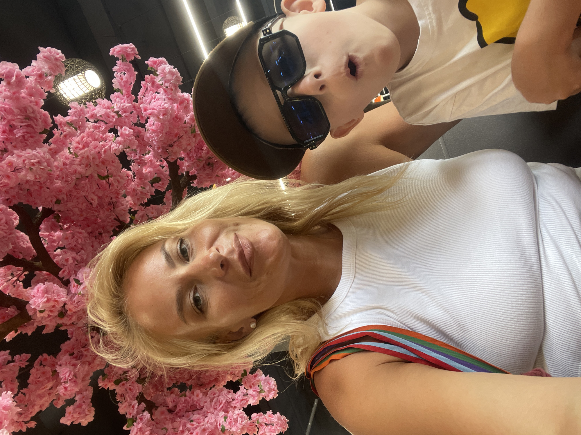 A medium shot captures a woman and a young boy posing for a selfie in what appears to be a restaurant or cafe. The woman, with blonde hair and wearing a white ribbed tank top, smiles at the camera. She has a pearl earring in her visible ear and a colorful striped strap is visible on her tank top. Beside her, the young boy, wearing a brown baseball cap and large dark sunglasses, looks at the camera with his mouth open in a surprised expression. He is wearing a white t-shirt with a yellow graphic. The background is dominated by an abundance of artificial pink cherry blossoms, suggesting a decorative interior. Overhead lighting fixtures are visible amongst the branches. The scene is set in Gagra, Abkhazia.