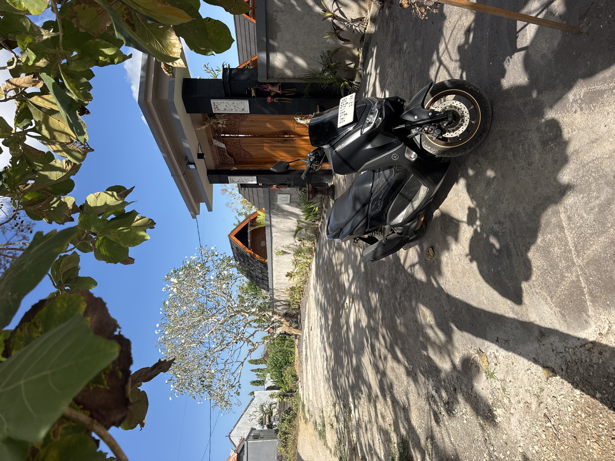 In Kuta Selatan, Indonesia, a black Yamaha NMAX scooter is parked on a gravel and concrete lot in front of a traditional Balinese-style house. The scooter's license plate reads "DK 3912 FDB". The house features ornate wooden double doors and a grey stone wall. A frangipani tree with white flowers is in bloom to the left of the house, and lush green foliage surrounds the property. The sky is clear and blue, and shadows from trees are cast across the ground. The overall scene depicts a sunny day in a tropical residential area.