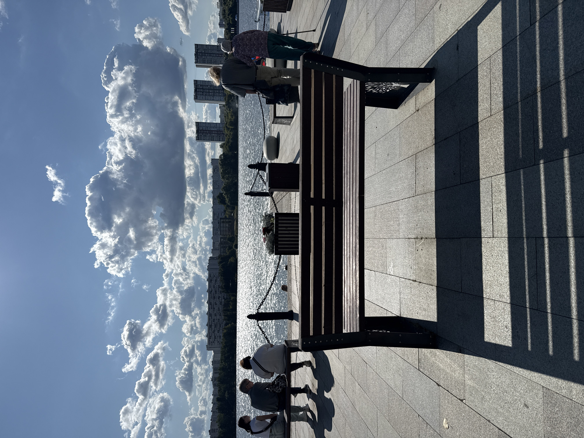 The image captures a sunny day by the Skhodnensky Bank of the Khimki Reservoir in Khimki, Russia. In the foreground, a dark wooden bench with patterned metal legs casts a long shadow on the grey tiled promenade. Beyond the bench, a group of four people are seated on another bench facing the water. Two individuals are standing further down the promenade, one appearing to lean on a cane. The reservoir is calm and reflects the bright blue sky filled with large, puffy white clouds. Across the water, the cityscape of Khimki is visible, featuring a line of apartment buildings and two taller, modern high-rise structures. The overall scene suggests a peaceful outdoor gathering on a pleasant day.