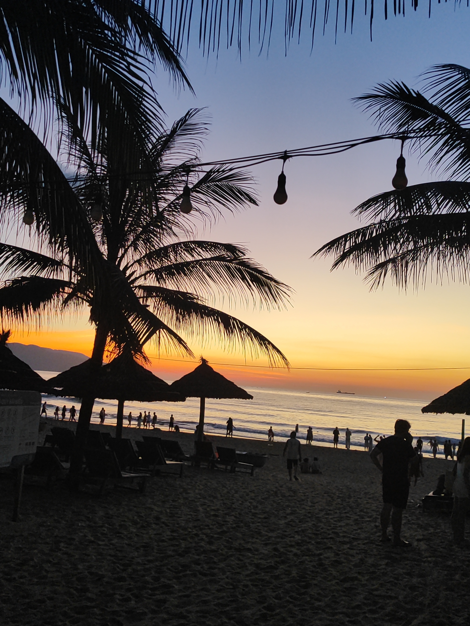 A sunset scene on a beach in Da Nang, Vietnam, features silhouetted palm trees with their fronds framing the top of the image. String lights with exposed bulbs hang between the trees. The sky displays a gradient of colors, transitioning from a soft purple and blue at the top to warm oranges and yellows near the horizon. The ocean is visible, with a few distant ships. On the beach, several thatched umbrellas stand, and rows of lounge chairs are set up. Numerous people are walking and standing on the sand, some near the water's edge and others further back. The sand itself appears dark and textured, suggesting it is late in the day. The overall atmosphere is peaceful and evocative of a tropical evening.