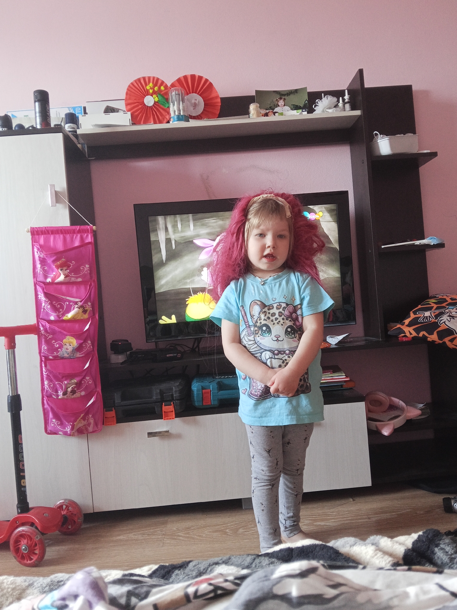 A young girl stands in a room in what appears to be a home setting. The room has pink walls. The girl is wearing a light blue t-shirt with a cartoon animal graphic on it, and gray leggings. She also has on a pink wig. Behind her, there is a TV set in a dark wooden entertainment center. On the shelves of the entertainment center are several decorations. To the left of the entertainment center is a tall white cabinet. A pink hanging organizer with images of Disney princesses is attached to the side of the cabinet. Next to the cabinet is a red scooter. A patterned blanket is on the floor in the foreground. This location may be in the city of Chaikovsky, Russia.