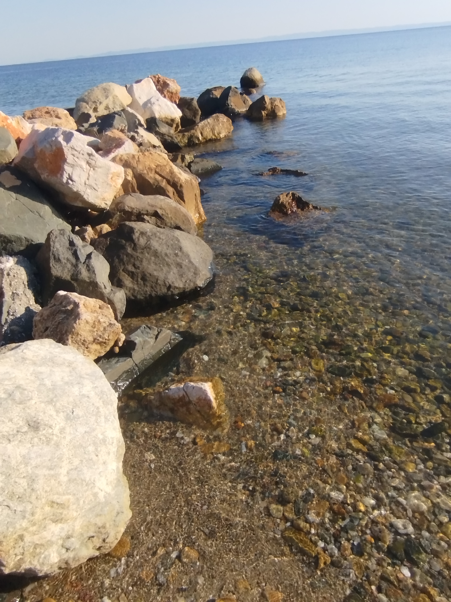 A body of water laps against a rocky shore on a clear, sunny day. The water is a deep blue in the distance, transitioning to a lighter, transparent blue closer to the shore. The seabed is visible through the clear water, revealing a bed of small, colorful pebbles and stones. A line of large, varied-colored rocks forms a barrier along the edge of the water, with some submerged and others rising above the surface. The rocks are a mix of light and dark grays, browns, and some with reddish-orange hues. The sky above is a pale, clear blue, with no clouds visible. The scene is a natural, serene coastal landscape, likely located in Sithonia Municipal Unit, Greece, given the context.