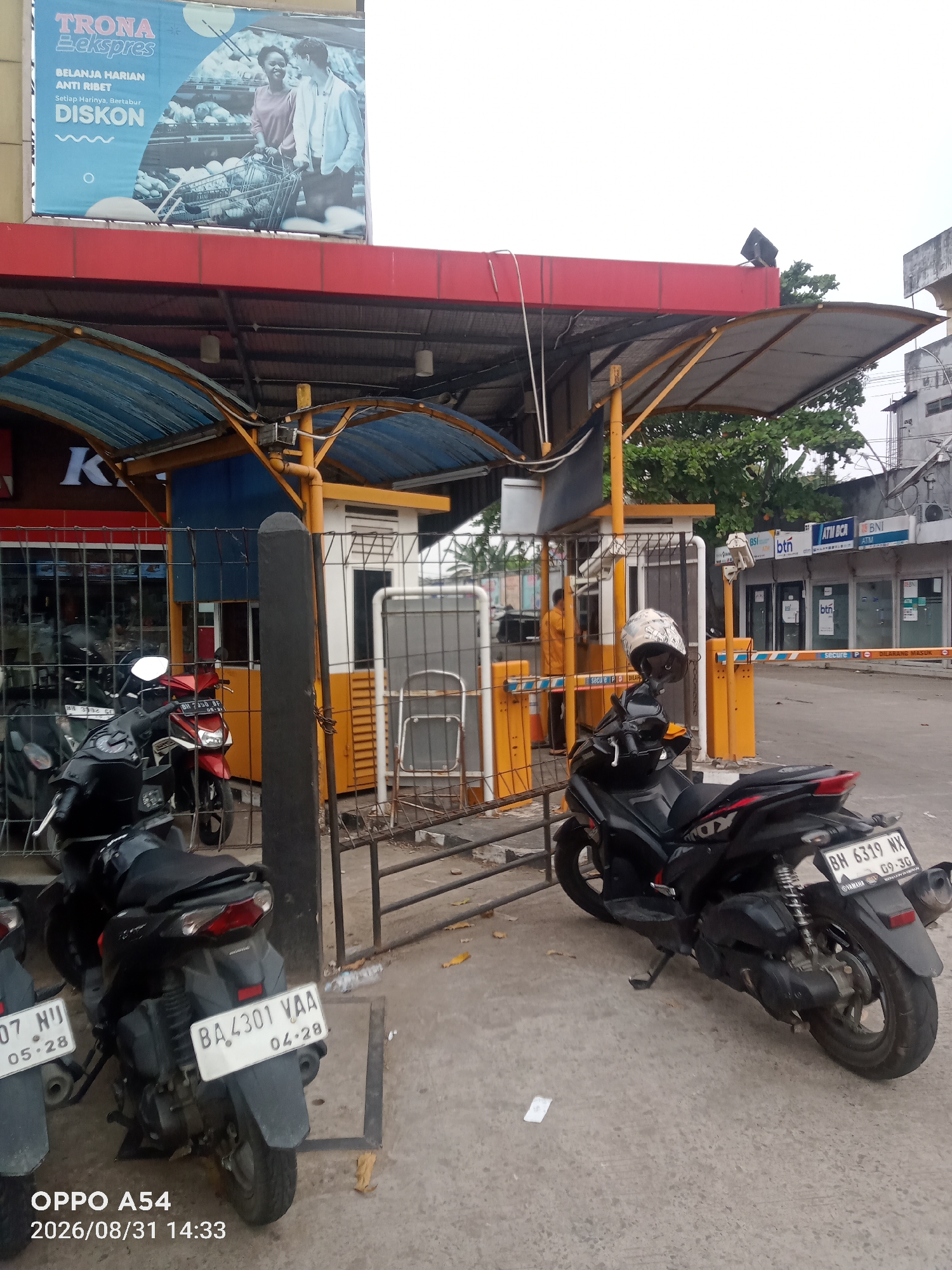 The image captures a street scene in Jambi, Indonesia, likely a parking area or entrance to a commercial establishment. In the foreground, several motorcycles are parked. One black motorcycle in the lower right has a license plate reading "BH 6319 NX". Another motorcycle to its left, partially visible, has a license plate "07 HIJ" and "05.28". Further to the left, a red motorcycle displays the license plate "BA 4301 VAA" and "04.28". A security booth with a yellow and white structure stands behind a metal fence. A person in an orange uniform is visible inside the booth. Above the booth, a large billboard advertises "TRONA Ekspres" with images of people shopping. The background shows buildings, trees, and a cloudy sky. The presence of ATMs from "BCA" and "BNI" suggests a commercial area. The overall environment appears to be an outdoor, urban setting.