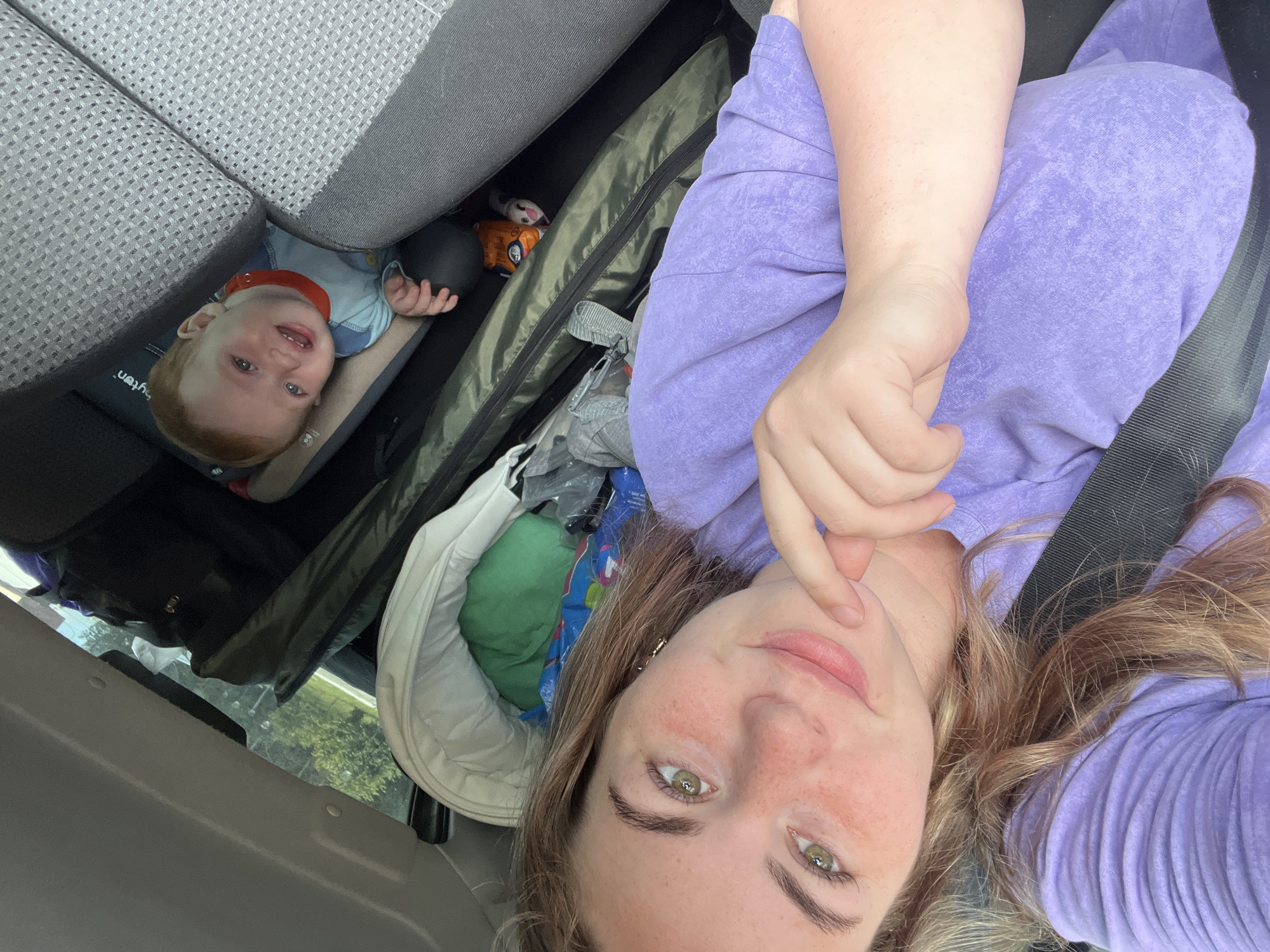 In a vehicle, a woman with blonde hair and freckles, wearing a purple t-shirt, takes a selfie. She has her hand near her chin and is looking at the camera with a slight smile. In the background, a baby is visible in a car seat, looking towards the camera with an open mouth, as if crying or speaking. There are several bags and items in the back of the vehicle. The scene appears to be in a car, possibly on a road trip, with trees visible through the window.