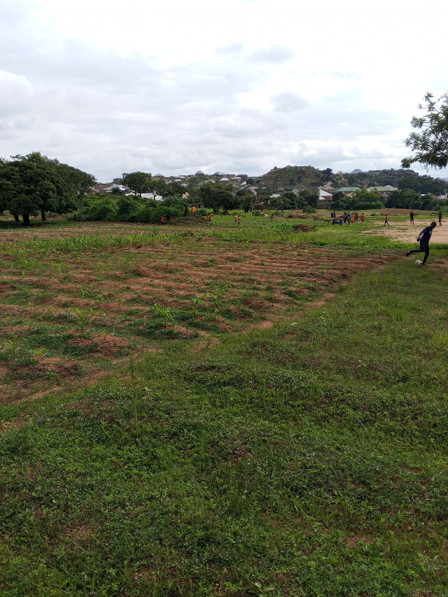 In Gwong, Nigeria, a group of children are playing soccer in an open field under a cloudy sky. The field is a mix of green grass and dry, reddish-brown soil with rows of young crops, possibly corn, planted in it. In the background, a village with various houses and buildings is nestled on a hilly terrain covered with trees. Some individuals, likely farm workers, are visible in orange clothing in the distance, tending to the crops. One child in a dark blue shirt is actively kicking a white soccer ball on the grassy area to the right, while another child in a similar shirt stands nearby. The overall scene depicts a rural setting where agricultural activities and recreational pursuits coexist.