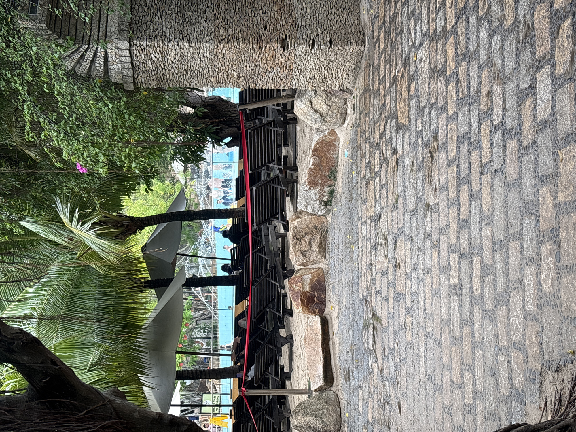 In Nha Trang, Vietnam, a resort scene unfolds with a view of a swimming pool area. The foreground is paved with cobblestones, with large rocks scattered on a sandy patch beyond. Rows of dark wooden lounge chairs are arranged under large white umbrellas, some unoccupied and some with individuals resting. A red rope cordon separates the lounge area from the pool. In the background, the bright blue water of the swimming pool is visible, with a water feature spraying water. A tall, stone-clad structure, possibly part of the resort's architecture, stands to the right. Lush green palm trees and other foliage frame the scene, with a single pink flower visible on a vine. A few people are casually dressed, suggesting a leisure setting.