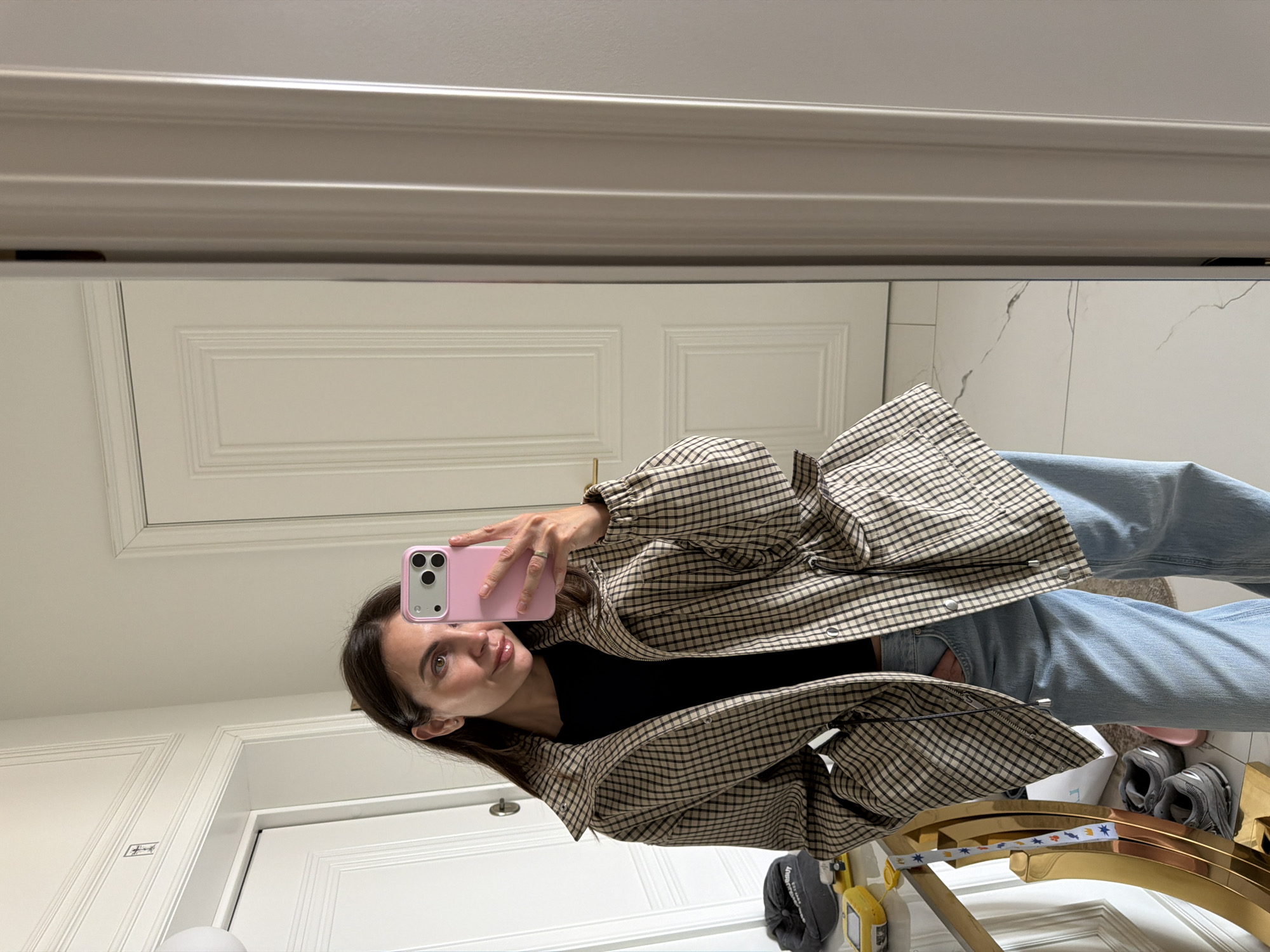 A person stands in a bathroom, taking a mirror selfie. The individual wears a checkered jacket, a black top, and light-wash jeans. A pink smartphone is visible in their hand. The bathroom features a white door with panel detailing, light marble floors, and a gold-colored decorative rack with personal items like shoes. The location is not identifiable as Krasnoyarsk, Russia from the provided image content.