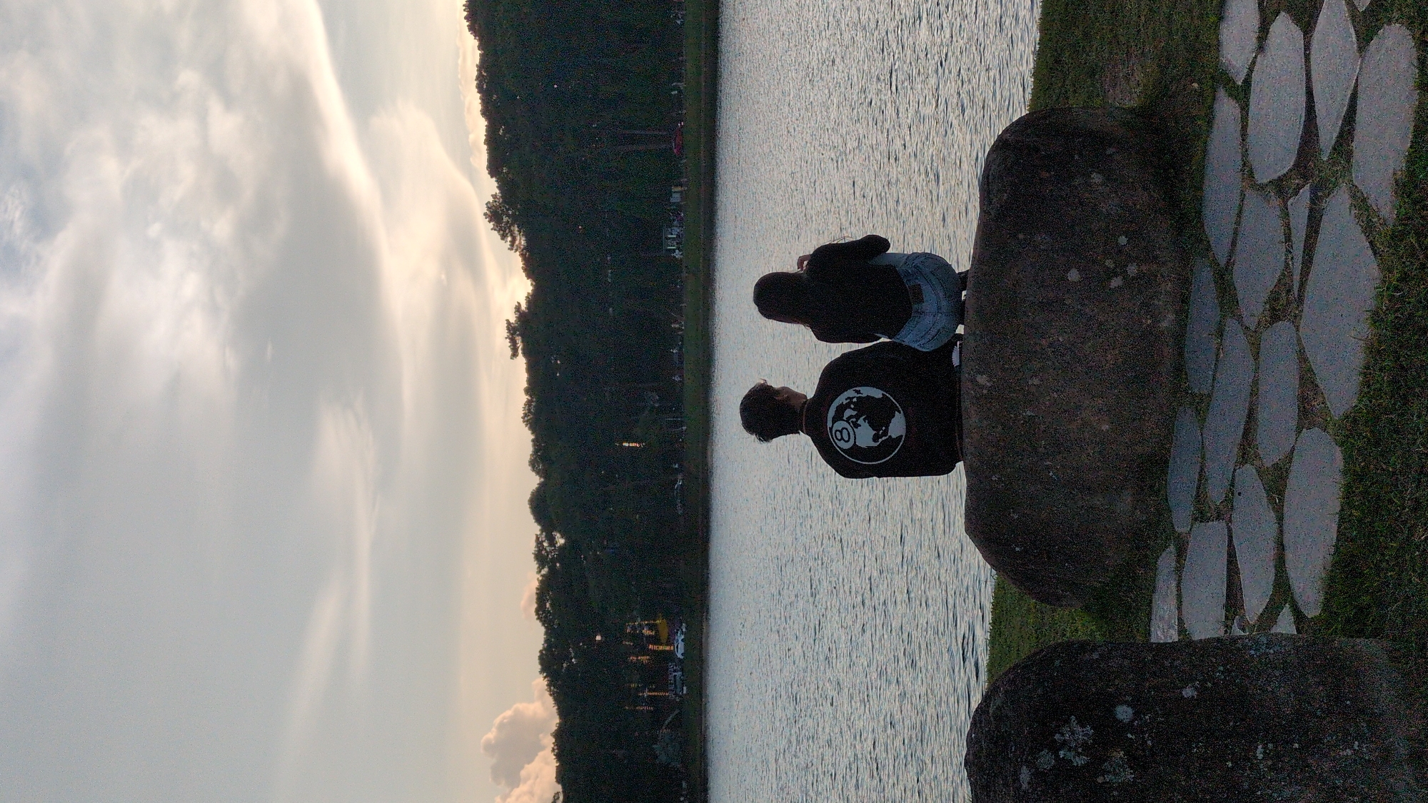 The image depicts two individuals, a male and a female, seated on a large rock overlooking a body of water, likely Xuan Huong Lake in Da Lat, Vietnam, given the scenery. The sky above is a muted blend of grey and pale yellow, suggesting either dawn or dusk, with wispy clouds. The lake's surface is rippled, reflecting the soft light. A dense line of dark green trees forms a backdrop along the far bank, with hints of distant buildings and lights visible through the foliage. The foreground features grassy terrain interspersed with irregular stone pathways. The male, on the left, wears a black jacket with a circular graphic featuring a globe and the number "8" on its back. The female, on the right, is dressed in a black top and light-colored denim shorts. Both are facing away from the camera, looking towards the lake. The overall atmosphere is serene and contemplative.