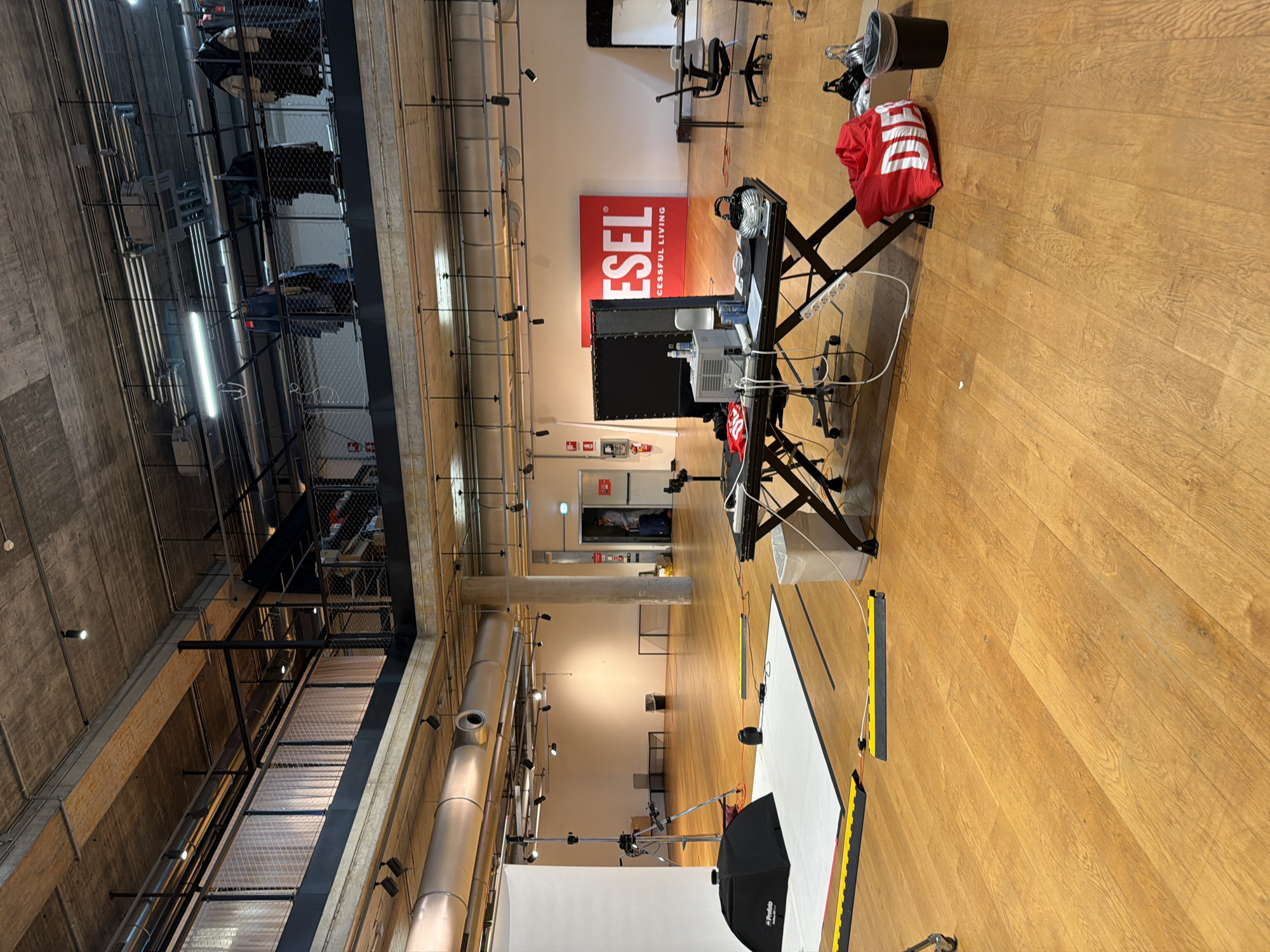 The image shows the interior of a large, open space, possibly a studio or warehouse location, in Mirabella, Italy. The floor is wooden and there is industrial-style overhead construction with metal walkways and exposed pipes. A red "DIESEL" logo is on a wall. There's a photography setup, including a backdrop, lighting equipment with an umbrella reflector, and a camera stand. A table holds electronic devices and a black screen is positioned nearby. A red bag and a black trash can are visible on the wooden floor. In the background are office chairs and a doorway with people visible inside.