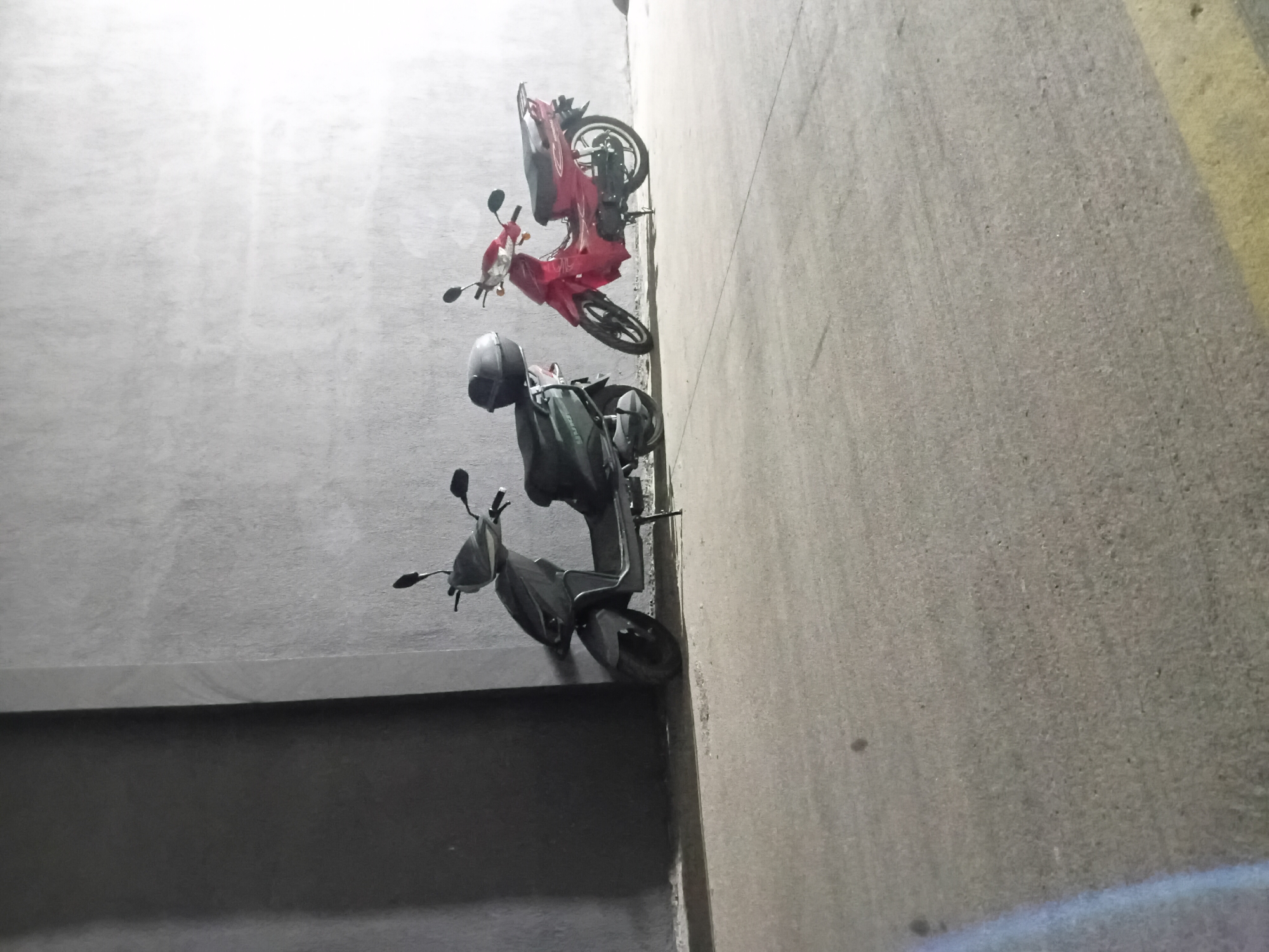Two scooters are parked on a paved surface next to a concrete wall in Onikişubat, Turkey. The scooter on the left is dark gray and appears to be an electric scooter. It has a black storage box on the rear seat. The scooter on the right is red and has a more traditional motorcycle design with exposed engine components. The background consists of a light gray concrete wall, with a darker vertical section on the left. The ground is a textured, light-colored concrete with some debris. The lighting suggests it may be evening or a dimly lit indoor space.