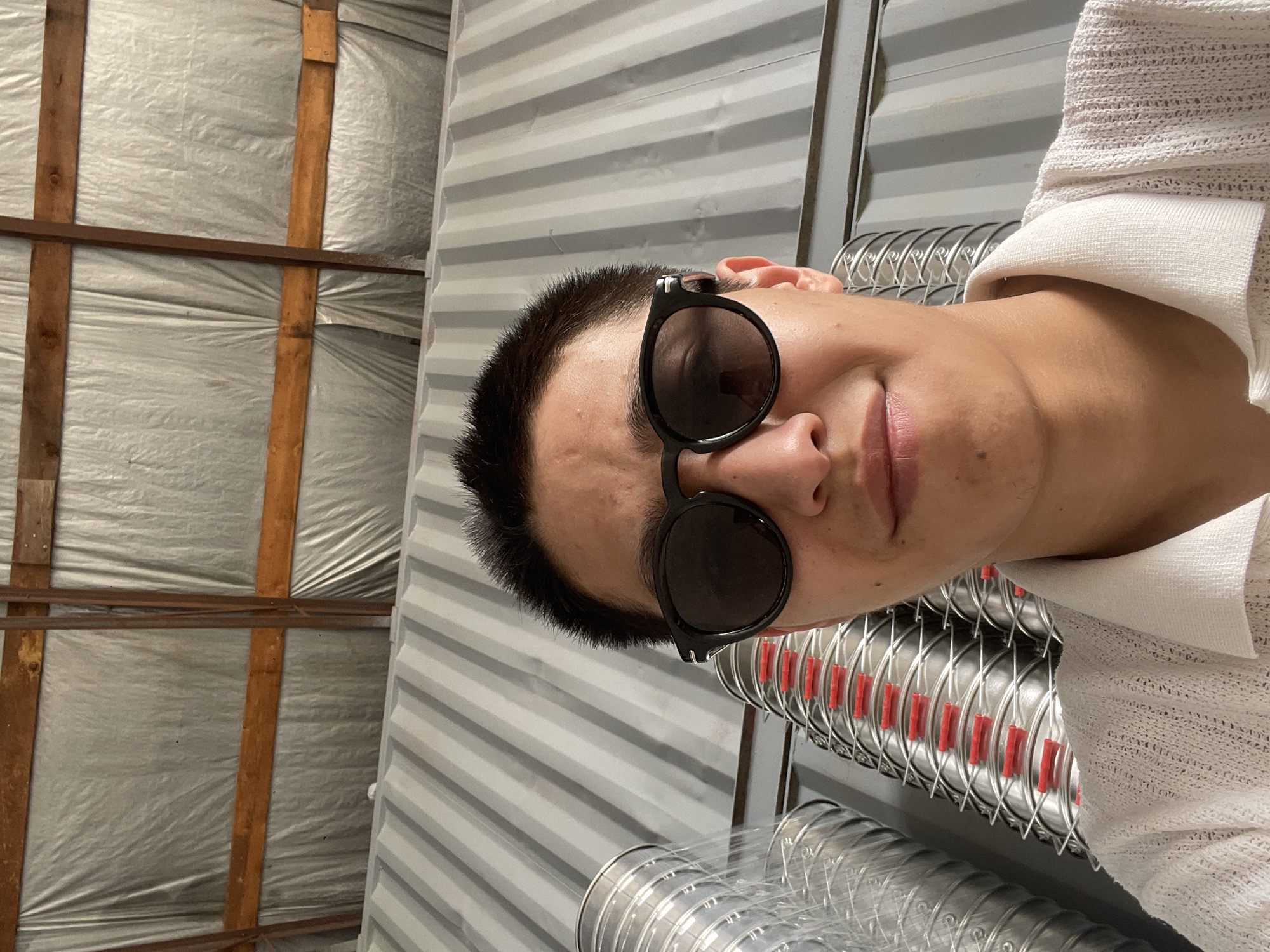A close-up, slightly upward-angled selfie shows a young East Asian person wearing dark, round-framed sunglasses and a light-colored, textured polo shirt. The person's short, dark hair is visible above the sunglasses. The background consists of corrugated metal walls and what appears to be insulated sheeting on a ceiling, with wooden beams supporting the structure. To the left and behind the person, stacks of cylindrical metal objects, possibly ductwork, with visible red plastic separators, are arranged vertically. The overall environment suggests an indoor industrial or storage space, likely in a workshop or warehouse setting, possibly located in the village of Kenen Azirbayev, Kazakhstan.