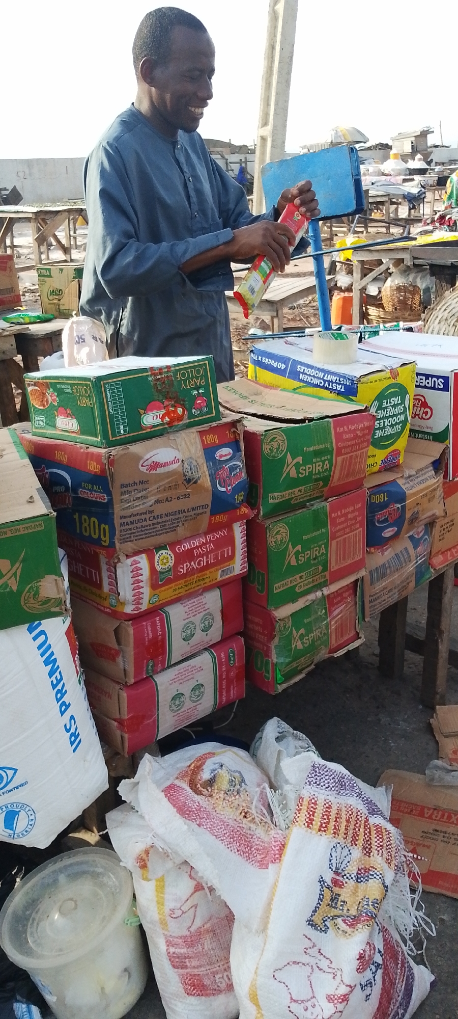 A middle-aged African man stands behind a makeshift stall laden with food products in Lokoja, Nigeria. He is light-skinned with short, dark hair and wears a blue, long-sleeved shirt. He smiles as he holds a red and green packet.
The stall displays stacks of boxed pasta and noodles. Brands like "Golden Penny Spaghetti," "Spira," "Supreme Noodles," "Party Jollof," "Milo" and "Alfamuda" are visible. White sacks, possibly containing rice, are placed on the ground near a clear plastic container. The immediate background shows more stalls and individuals, creating a market scene. Further in the background, there are structures, suggesting a city setting. A blue object is above the goods on sale on a pole.