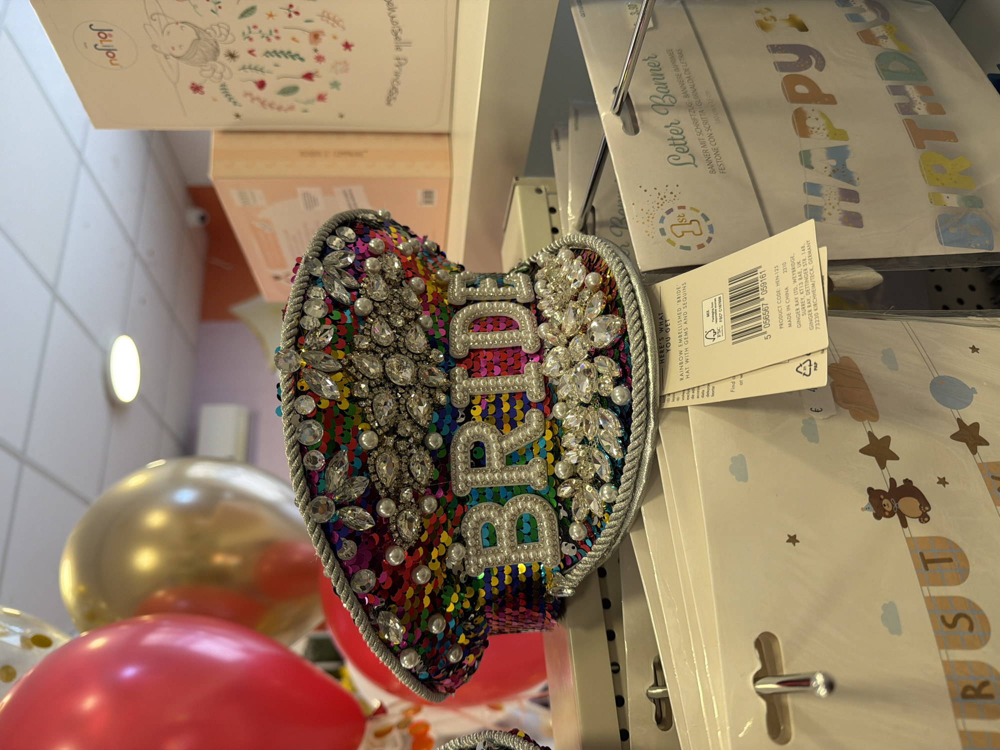 The image depicts a close-up of retail items, specifically a sequined heart-shaped "Bride" hat with gems and pearls, hanging on a display rack in a store. Beside the hat, other products are visible, including a "Letter Banner" package, likely for a birthday, and other party-themed items. Balloons are visible in the background. No information about the specific location in Budva, Montenegro is available in the image.