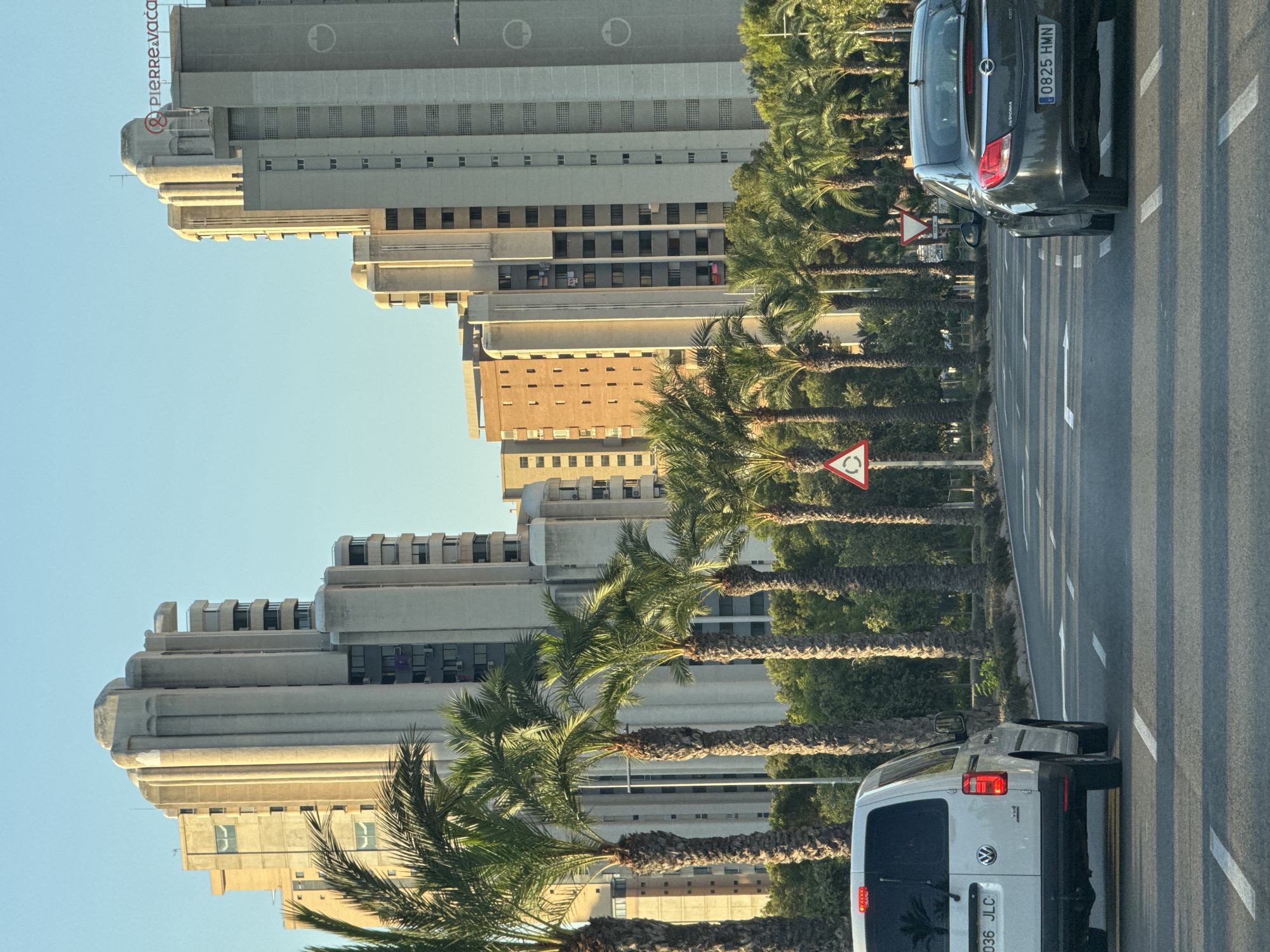 The image shows a street scene in Benidorm, Spain, with tall, modern buildings and palm trees lining the road. The sun is setting or rising, casting a warm glow on the buildings. Two cars are visible on the road: a white van on the left and a dark-colored sedan on the right. Both cars are driving on the right side of the road. A traffic sign indicating a roundabout is visible on the left side of the road. The buildings appear to be residential or hotel complexes, with balconies and windows. The sky is clear and blue. The overall impression is of a bright, sunny day in a busy urban environment.