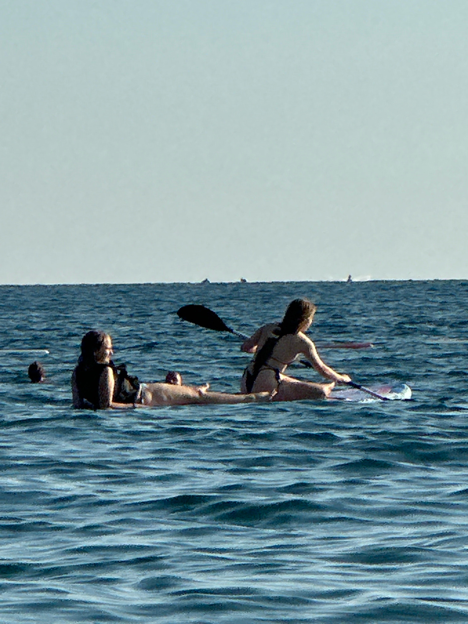 Two women are on a paddleboard in the sea, with the city of Boreti, Montenegro visible in the distance. The woman in the foreground is lying on her stomach, facing to the right, wearing a black swimsuit and a black life vest. The second woman is sitting up on the paddleboard, facing to the left, wearing a black bikini and holding a paddle. The water is choppy and blue, with a clear sky above. There are a few other distant objects on the water that appear to be boats. The scene appears to be a sunny day.