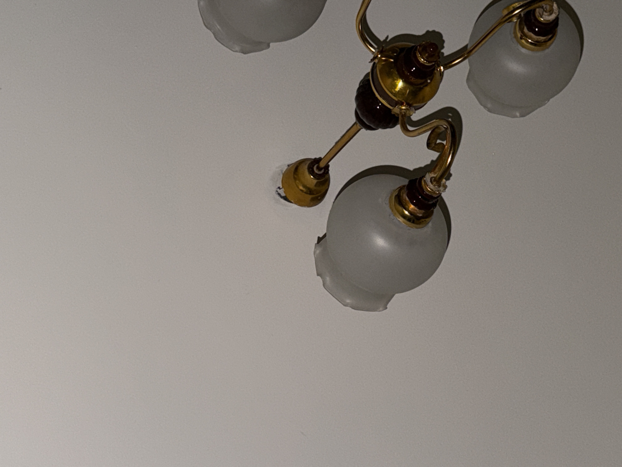 The image shows a close-up, angled view of a ceiling light fixture against a plain, light-colored ceiling. The fixture is made of brass with dark brown accents and frosted glass shades. At least three shades are visible, with one shade on the left appearing chipped or broken. One of the light sockets, on the upper left, is exposed and appears to be missing a light bulb. The fixture is positioned in the upper right portion of the frame, with the majority of the image space occupied by the ceiling. The lighting is dim, casting subtle shadows. There are no people or other objects visible in the frame. The location is stated to be in the city of Khimki, Russia.