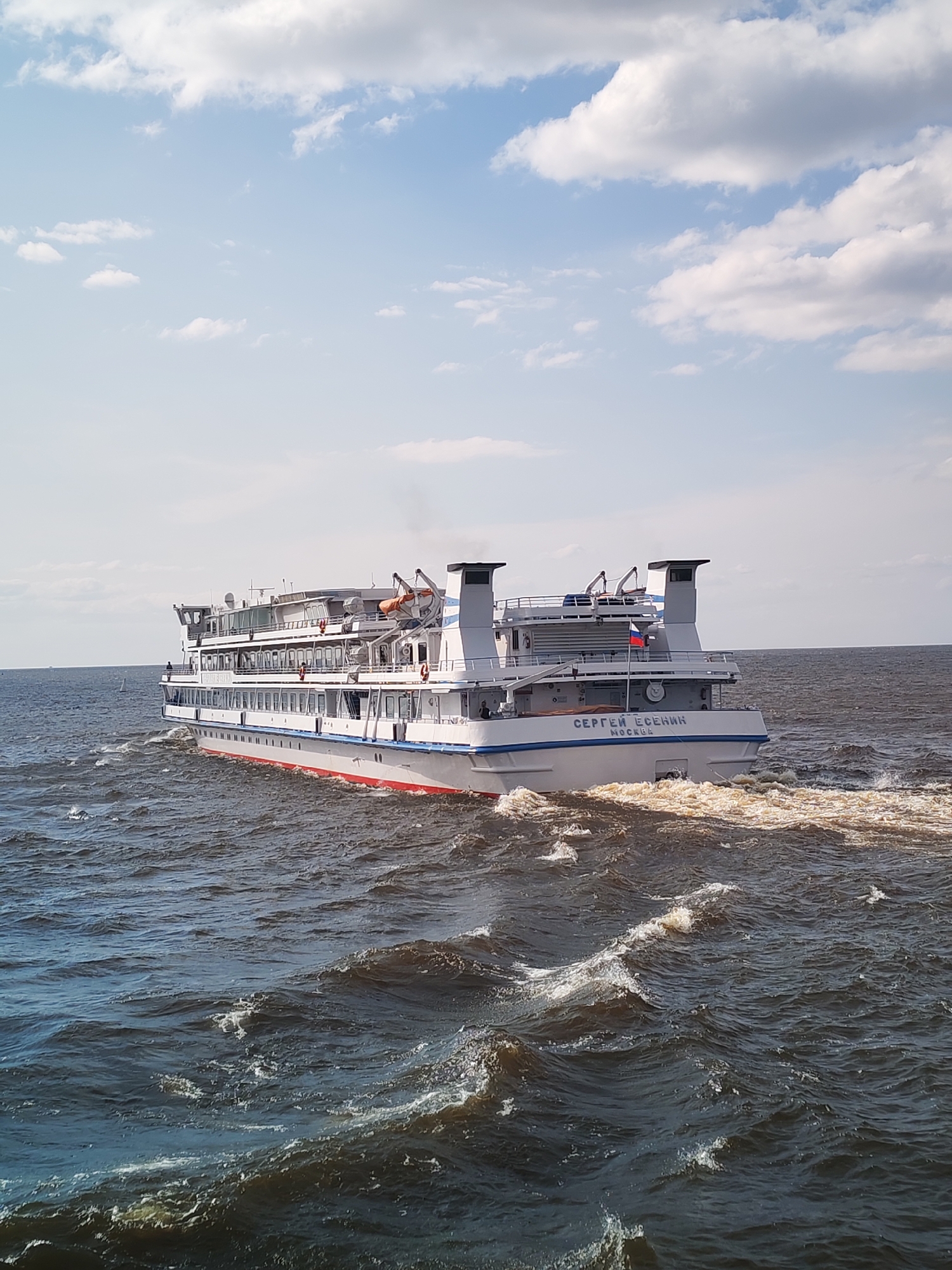 In the city of Krokhino, Russia, a large white passenger ship named "SERGEI YESENIN MOSCOW" sails on a dark, choppy body of water under a partly cloudy blue sky. The ship has multiple decks with numerous windows and is creating a visible wake. The ship's hull is white with a red stripe near the waterline, and it has two prominent structures on its upper deck that appear to be smokestacks or exhaust vents. A Russian flag is visible on the upper deck. The water is dark blue and shows significant wave action, with white foam and spray visible around the ship's stern. The horizon is visible in the distance, with a faint line separating the water from the sky. The overall scene depicts a ship in motion on a somewhat rough body of water.