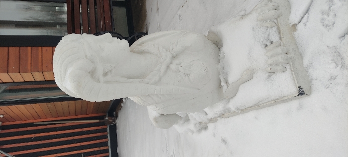 An outdoor scene in Grebnevo, Russia, displays a white sculpture heavily covered in fresh, powdery snow, giving it the appearance of a snow sculpture. The central subject is the bust of a female figure, depicted in profile facing right, with prominent ram-like horns or a stylized headdress extending backward from her temples. Her hair or headdress features horizontal ridges, and a textured, possibly braided, necklace is visible around her neck. The figure rests on a rectangular, snow-covered base that includes stylized clawed paws at its front. The entire ground surface is uniformly covered in a layer of white snow. In the background, a building with vertical reddish-brown wooden siding is visible. A tall, narrow dark-framed window reflects the sky, and to its left, a dark vertical structural element is present. A long-handled shovel with a light-colored blade leans against the building wall to the far left. To the right of the sculpture, a dark brown wooden slatted bench is partially visible against the building. The scene is static, illuminated by ambient daylight consistent with a winter day.